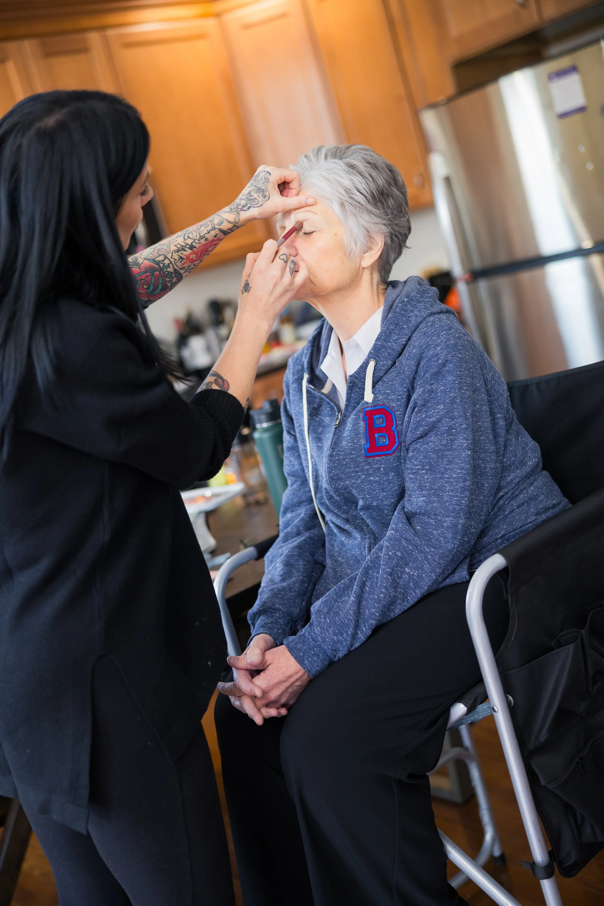 A young woman with tattoos applying makeup to an elderly woman with gray hair, who is sitting in a wheelchair in a kitchen.