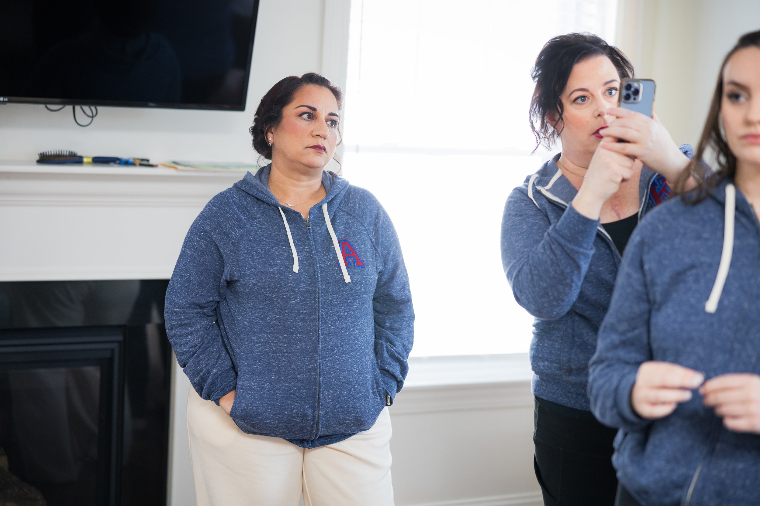 Three women in casual attire, two of them are wearing matching blue hooded sweatshirts, one with a large letter A on the chest, standing indoors with natural light, with a large black TV mounted on the wall and some tools on a white shelf in the back