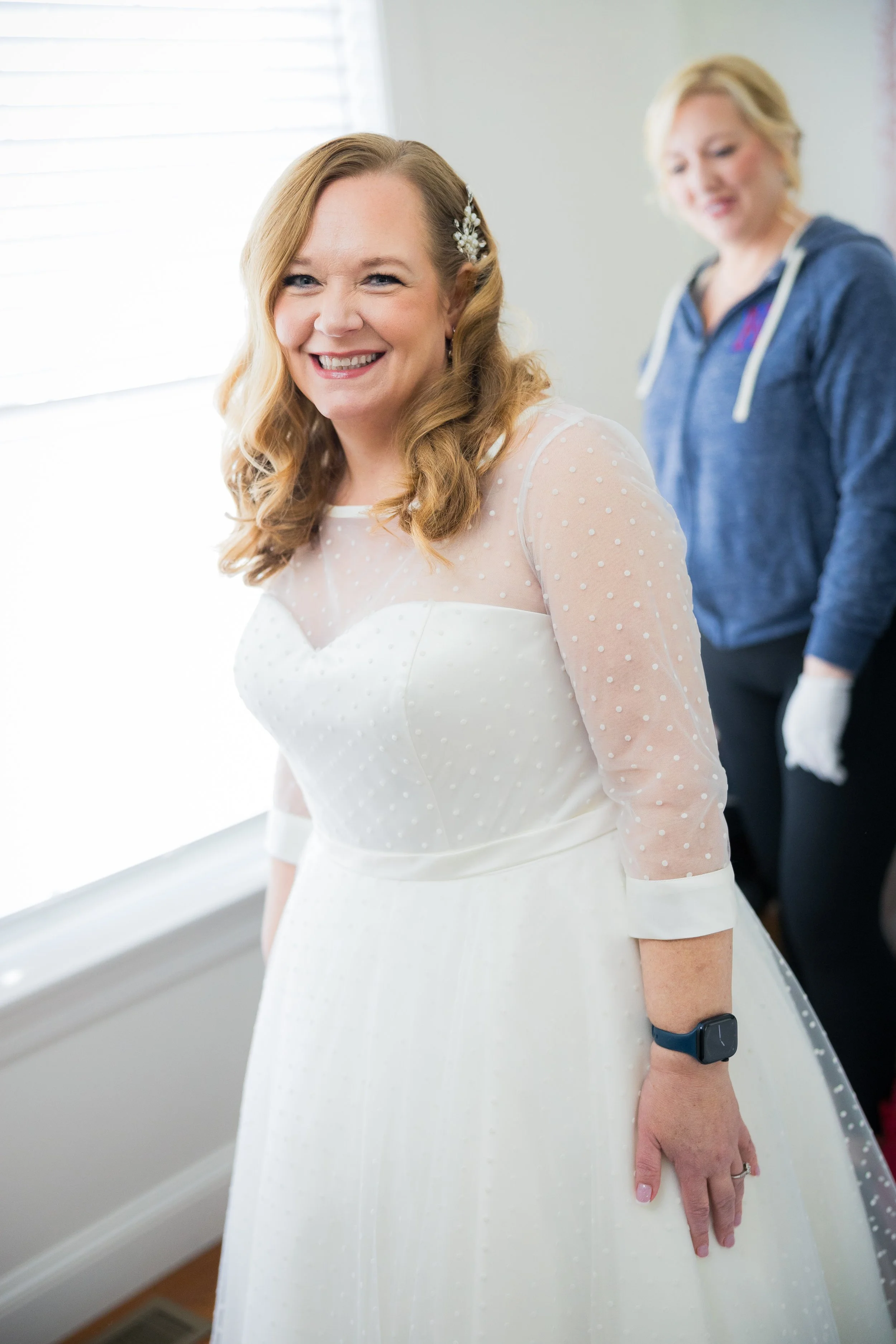 A smiling woman in a white dress with sheer, polka-dotted sleeves, standing by a window with white blinds. Another woman in a blue hoodie is behind her, slightly out of focus.