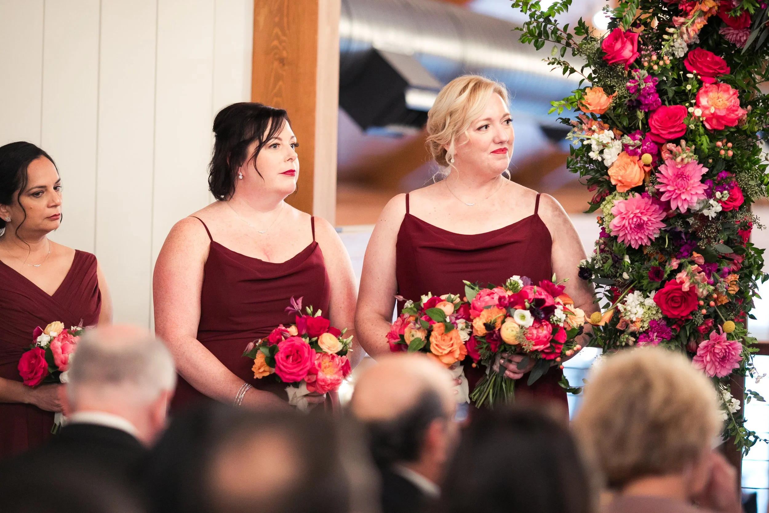 Women in burgundy dresses standing next to each other at a gathering with large floral arrangements of pink, orange, white, and purple flowers.