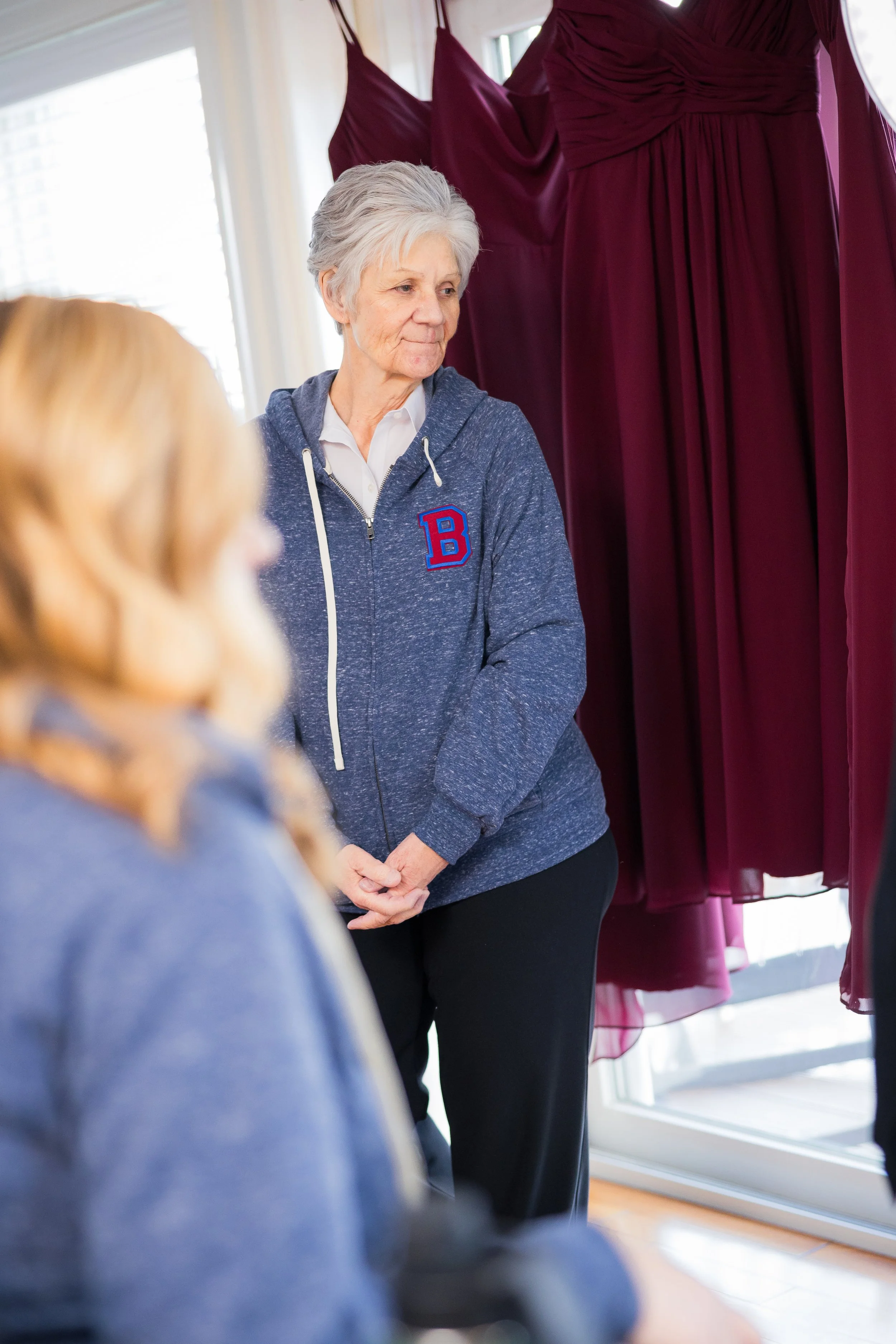 An elderly woman with gray hair wearing a blue hoodie with a red and blue letter 'B' on it, standing indoors in front of a window with maroon curtains, and a younger person with blonde hair blurred in the foreground.