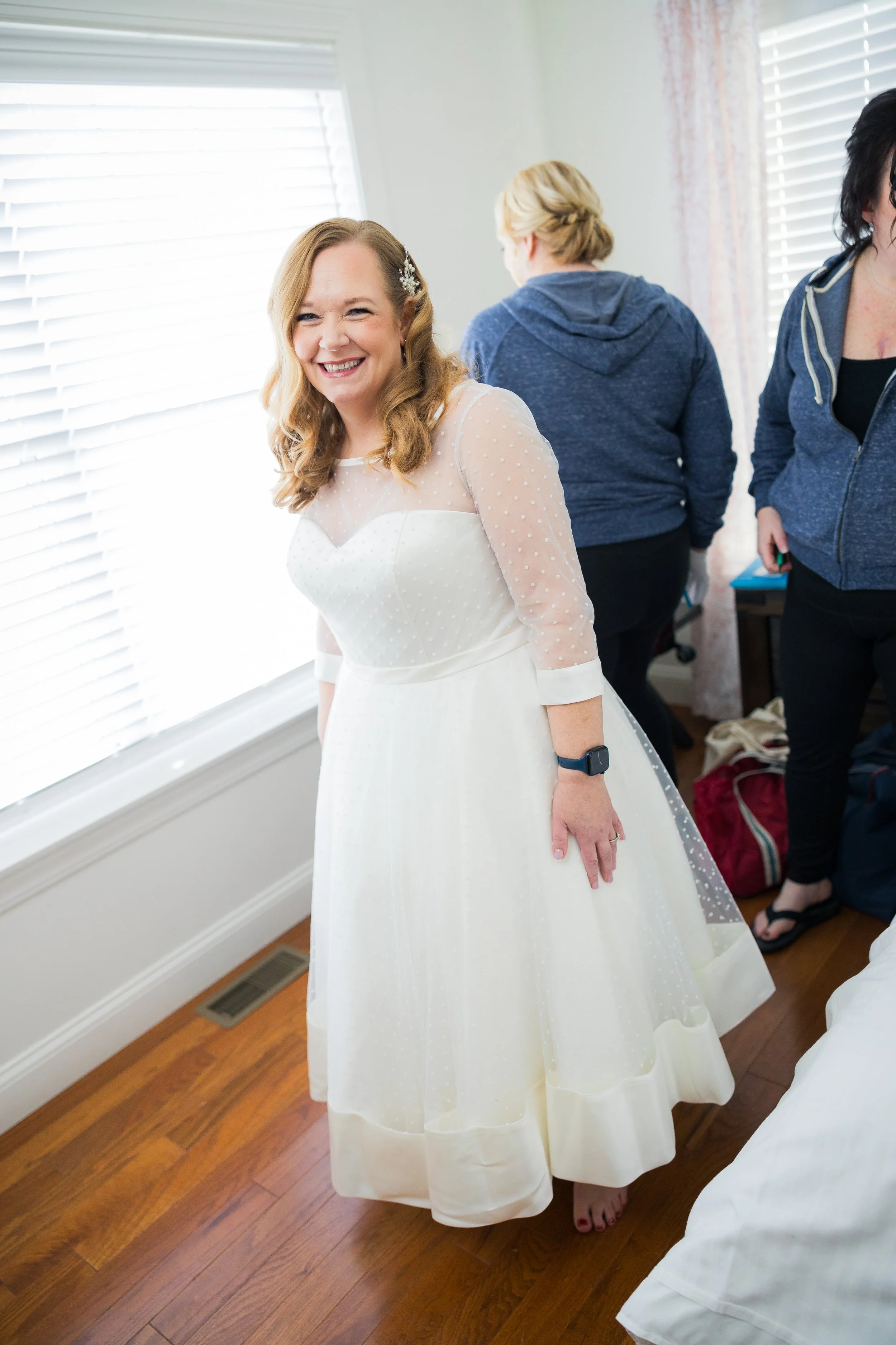 A smiling woman in a white wedding dress standing near a window, with two women in casual clothes in the background.