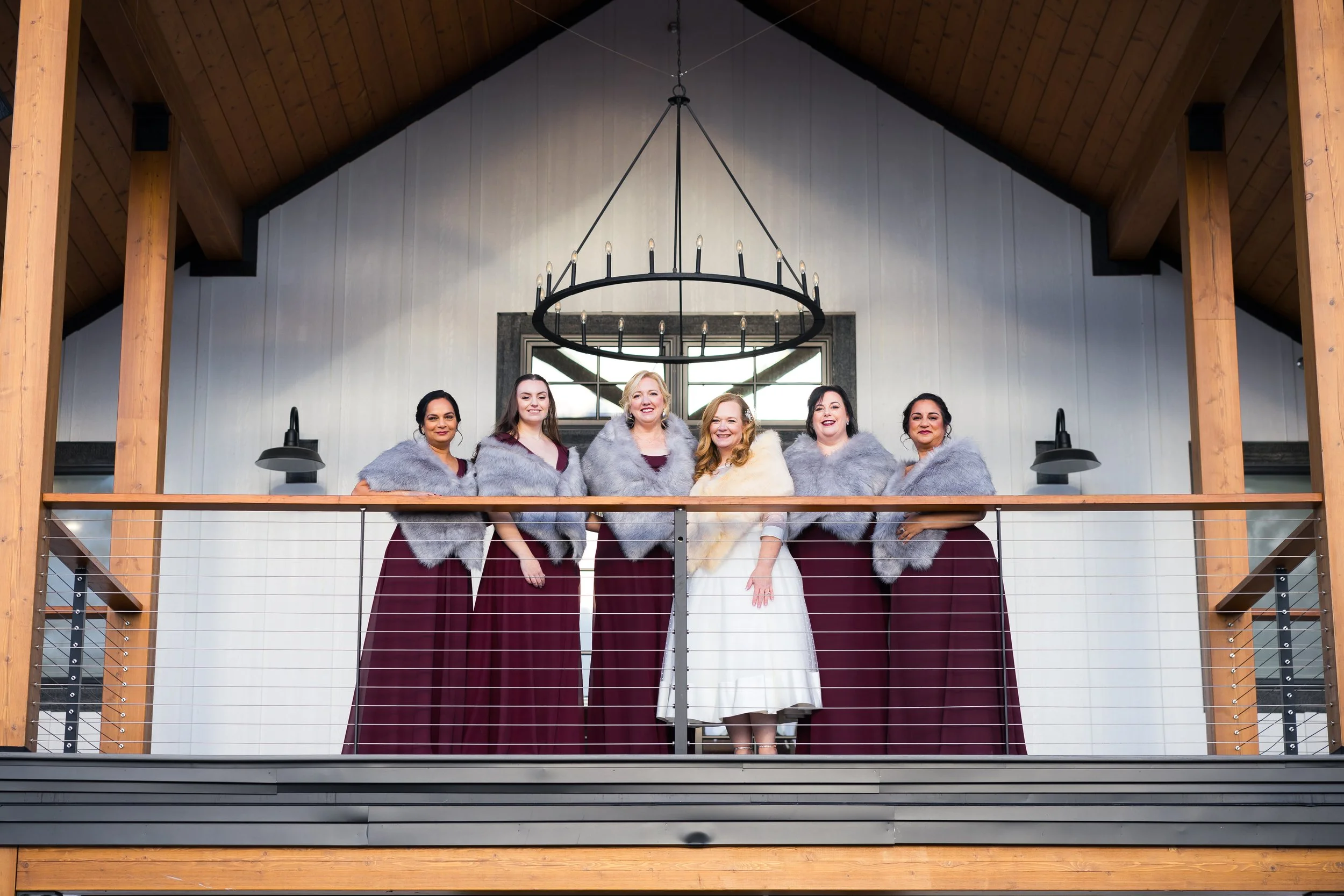 A wedding party with six women standing on an indoor balcony, dressed in formal attire and furry shawls, with a wooden ceiling, chandelier, and large window behind them.
