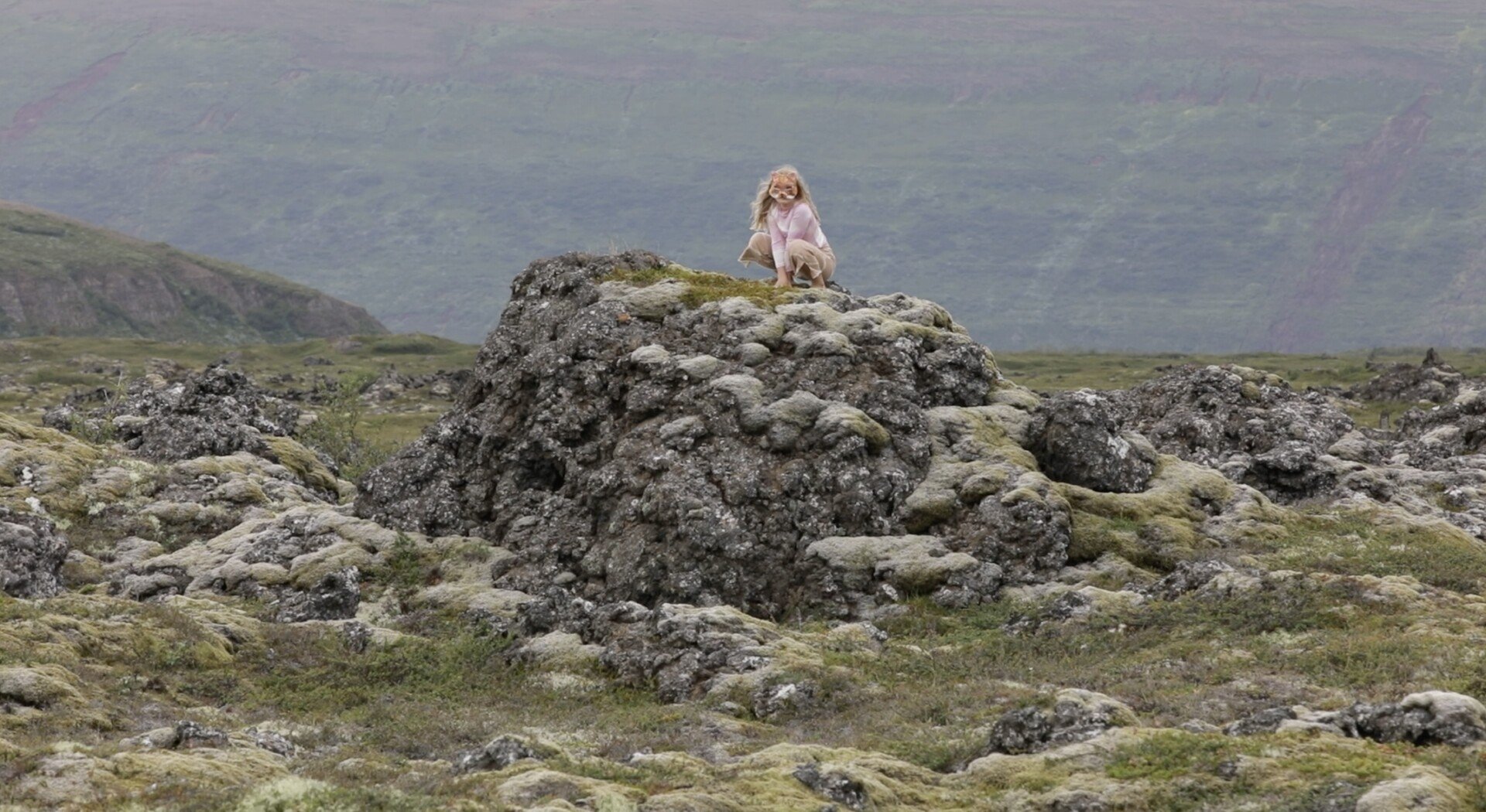 A woman crouched on top of a large mossy rock in a vast, rocky, and moss-covered landscape with mountains in the background.
