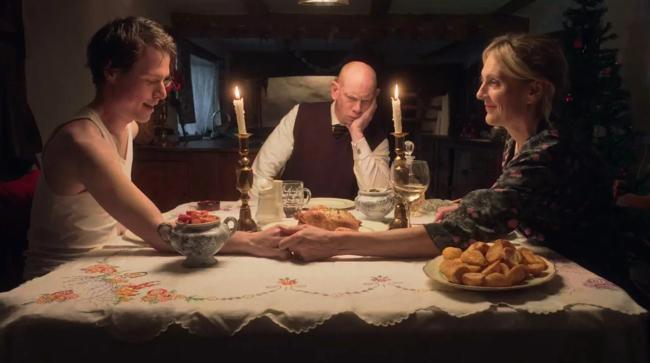 Three people sitting at a decorated dinner table in a cozy room, holding hands and bowing their heads in prayer. There are lit candles, a turkey, and various side dishes on the table, suggesting a holiday meal celebration.