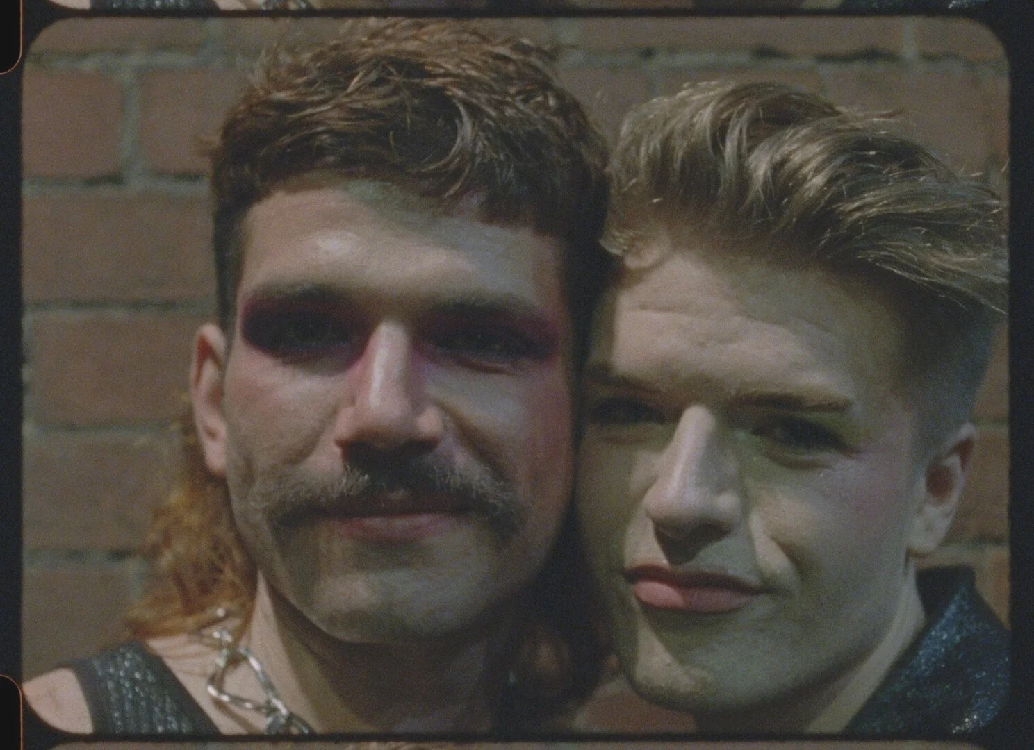 A close-up photograph of two people with makeup and distinct styles standing against a brick wall. The person on the left has dark eye makeup, blush, a mustache, and wavy hair. They are wearing a chain necklace and a sleeveless top. The person on the right has greenish makeup on their face, styled hair, and is wearing a black garment with a shimmer. The photo appears to be moody and artistic.