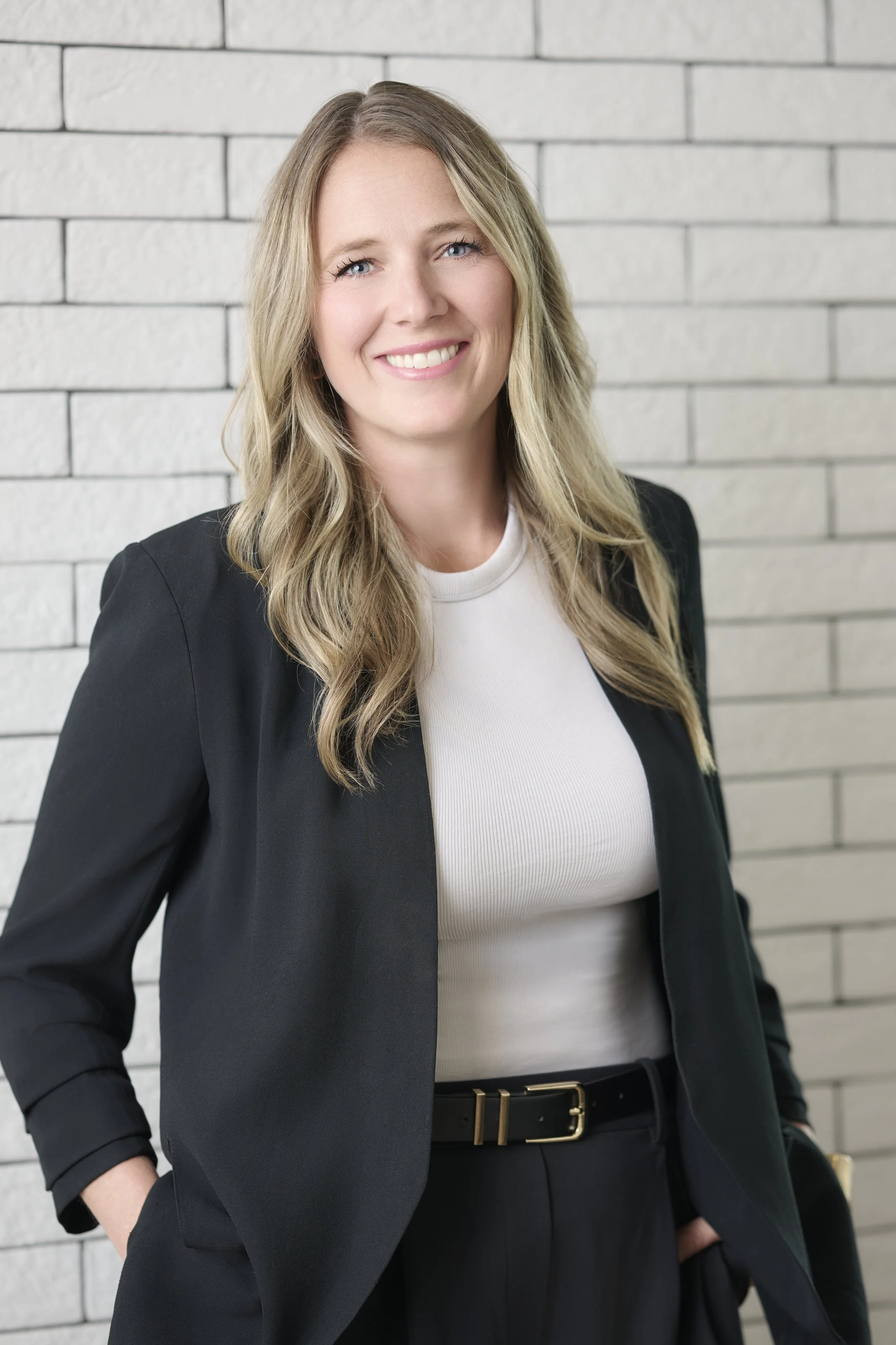 Woman smiling, seated in a white office chair in a modern office space with white brick wall and large windows.