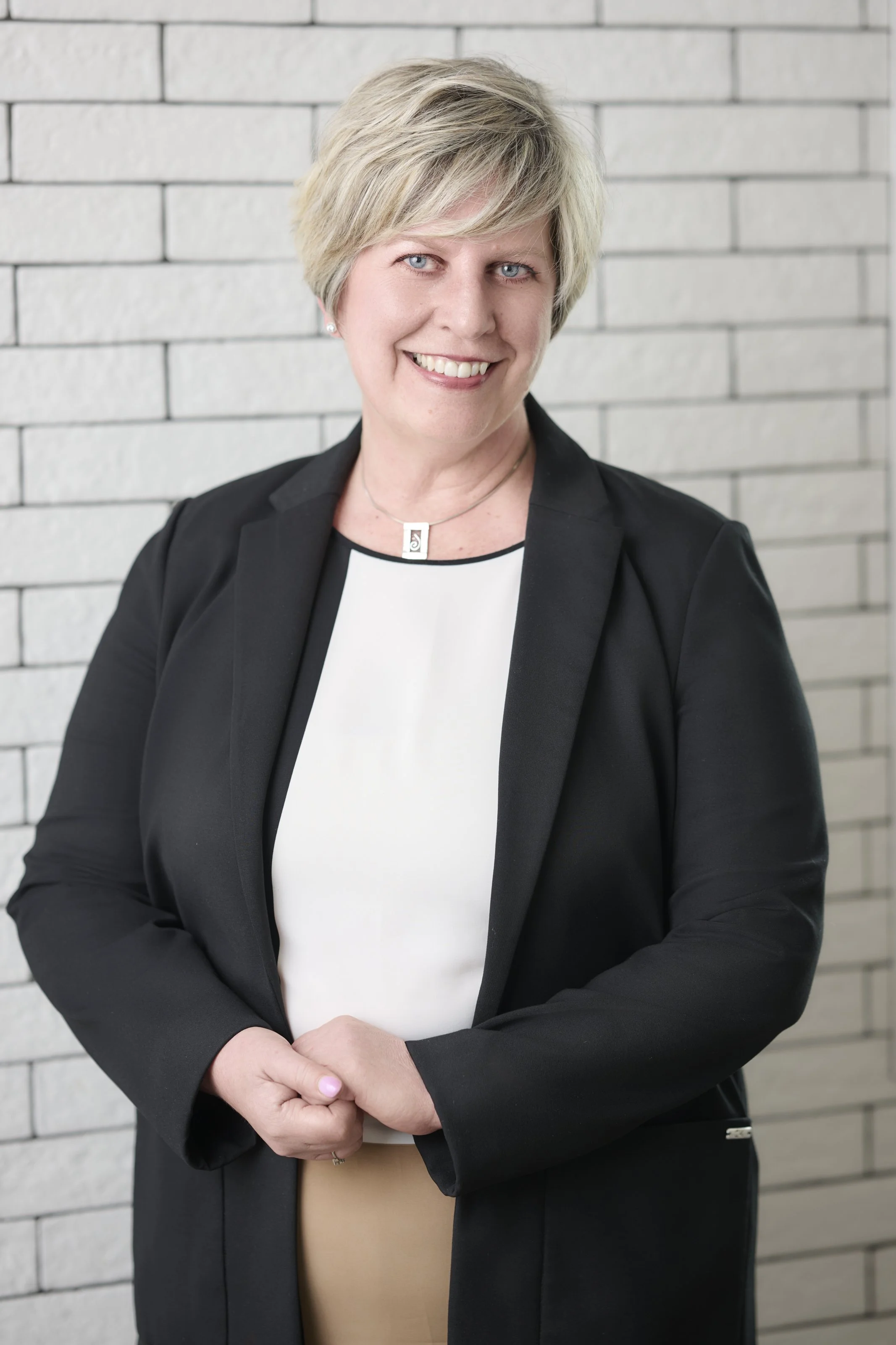 A woman with short blonde hair smiling, wearing a black blazer over a patterned blouse, standing near a wall with windows in the background.