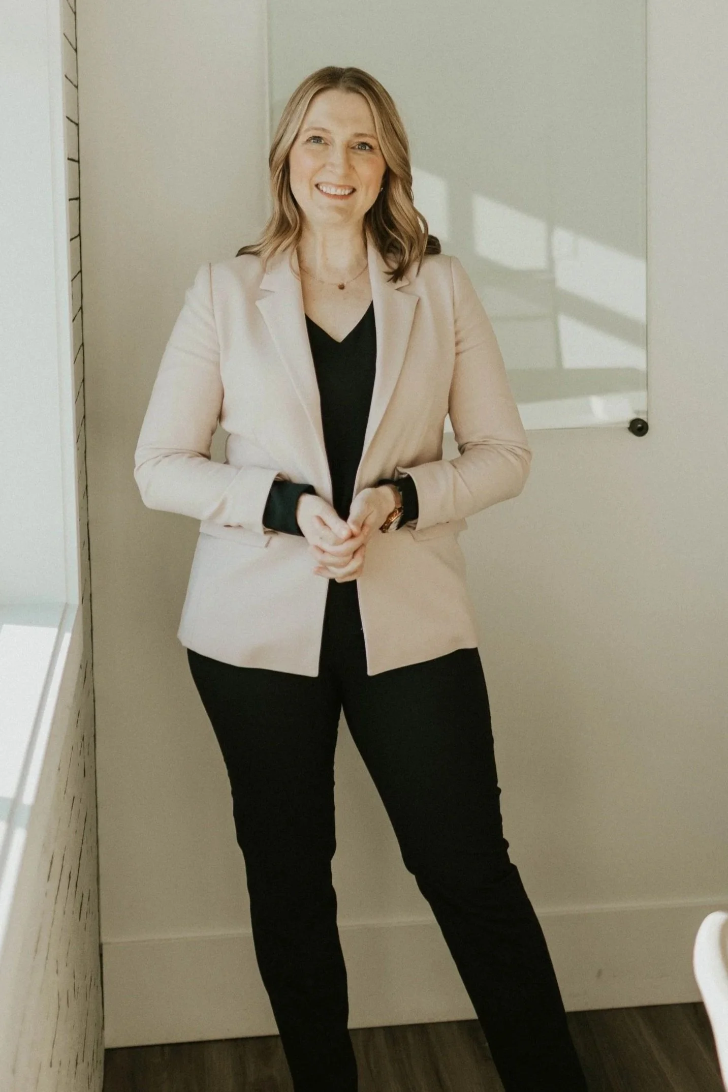 A woman with shoulder-length blonde hair smiling and standing in an office with a whiteboard behind her.