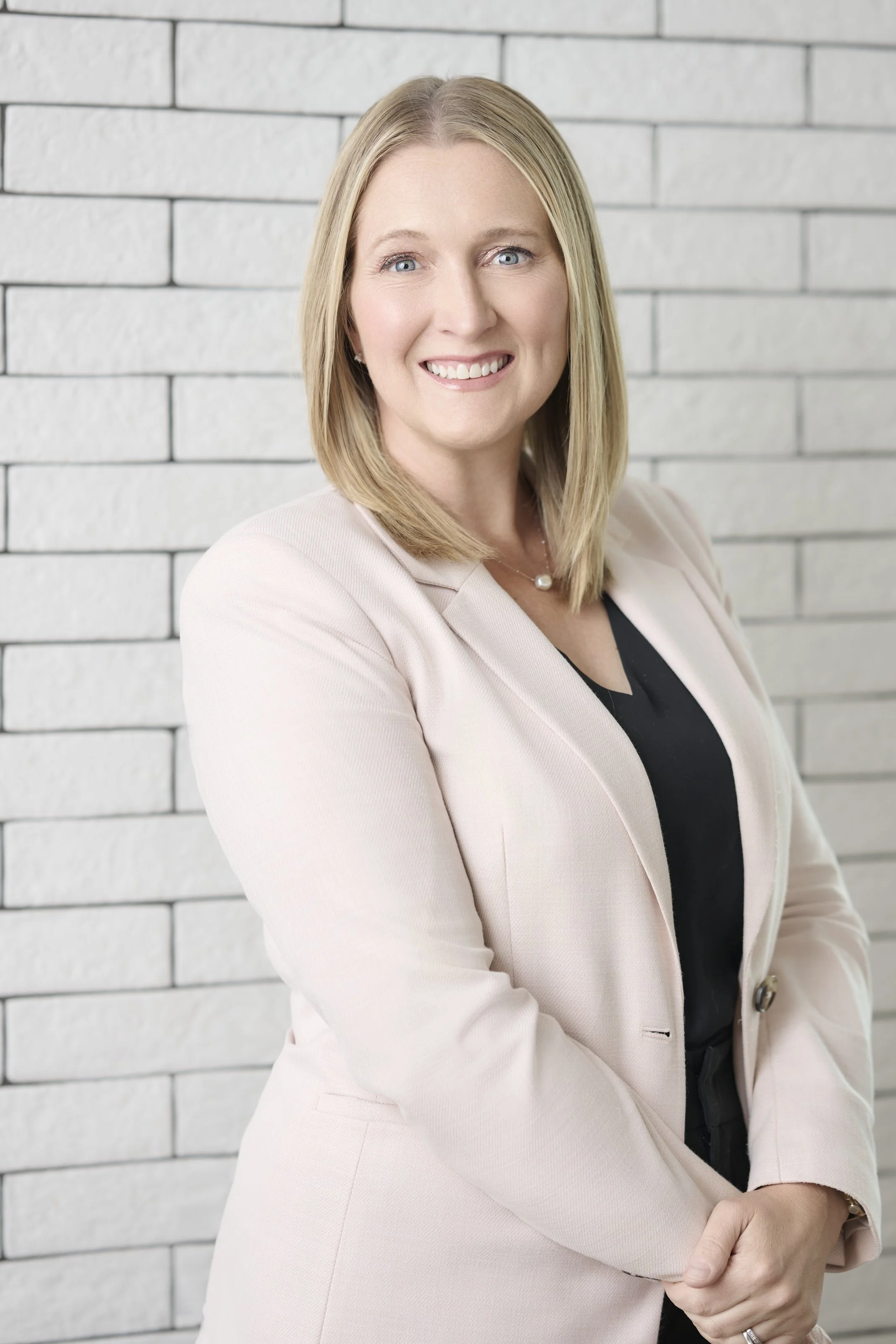 A woman sitting in a white office chair smiling, wearing a black top, beige blazer, and black pants, in a modern office with white walls, brick accent wall, large windows, and a white table.