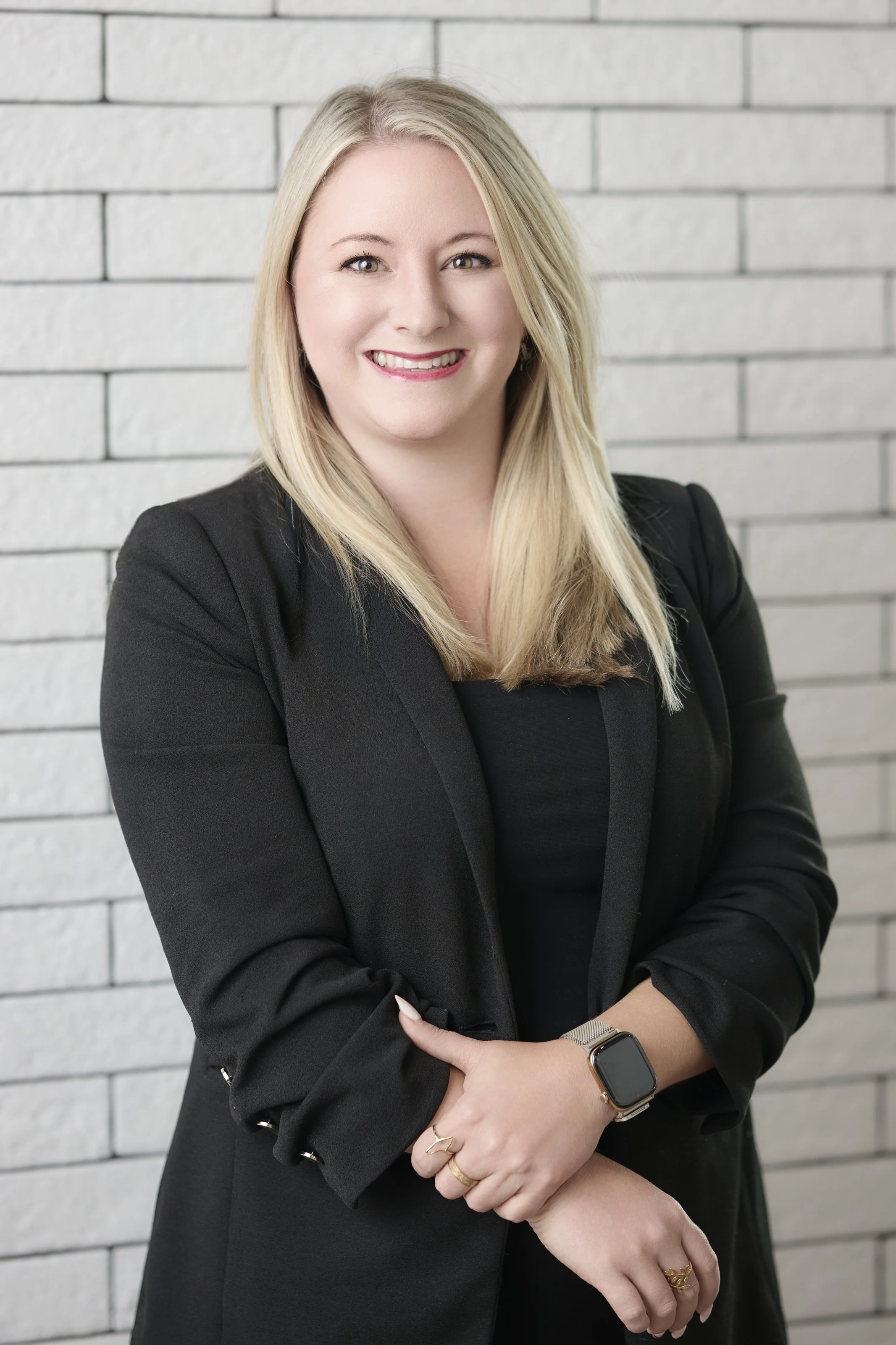 A woman with long blonde hair, smiling, standing with arms crossed in front of a white brick wall, wearing a red blazer and black top.