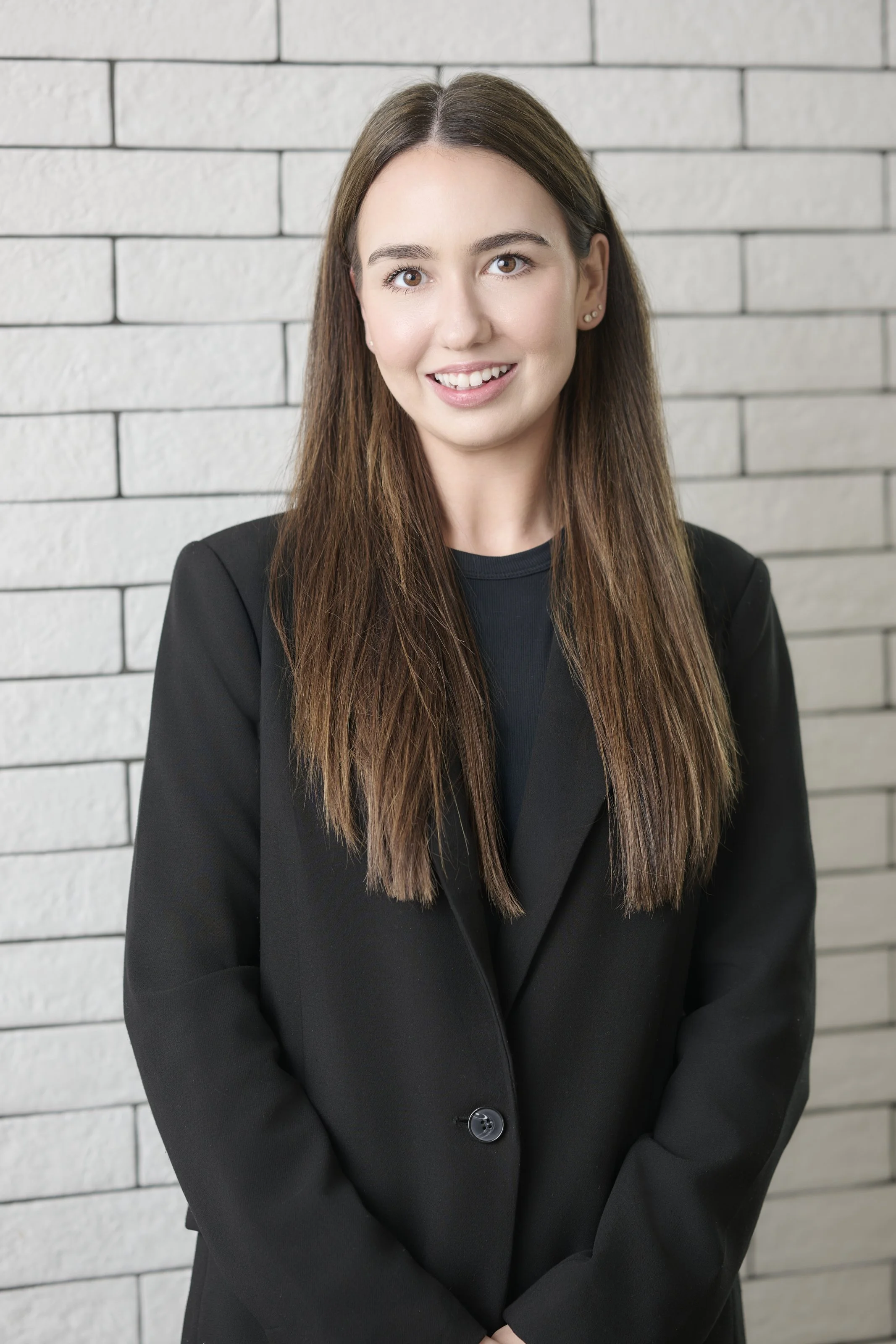 A woman with long, wavy brown hair, wearing a black blazer, standing in front of a white tiled wall.