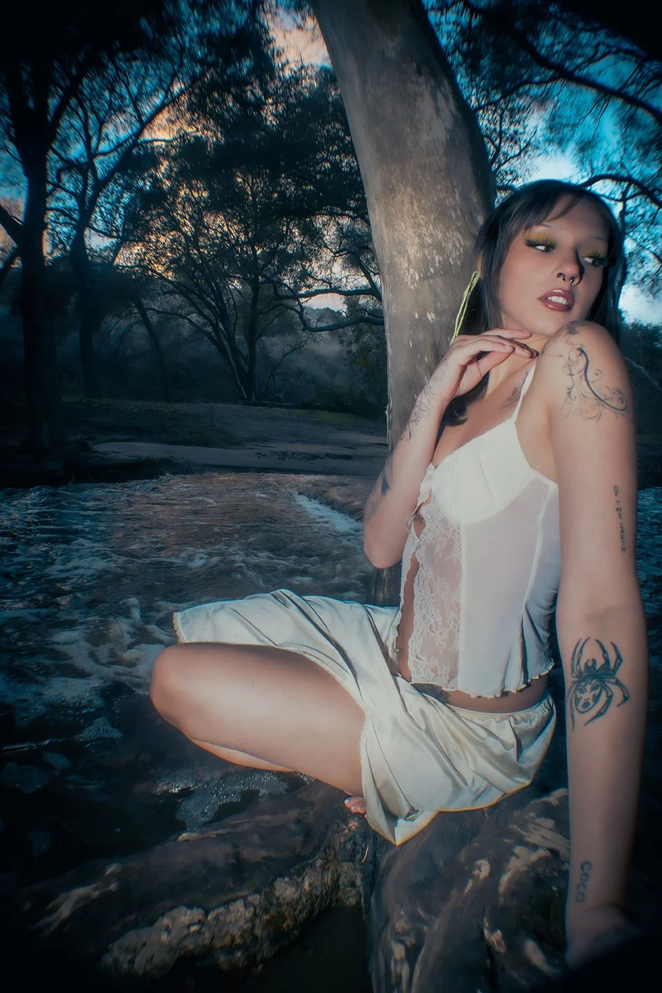 Young woman with tattoos sitting in a shallow stream near a large tree trunk, with trees and a sunset sky in the background.