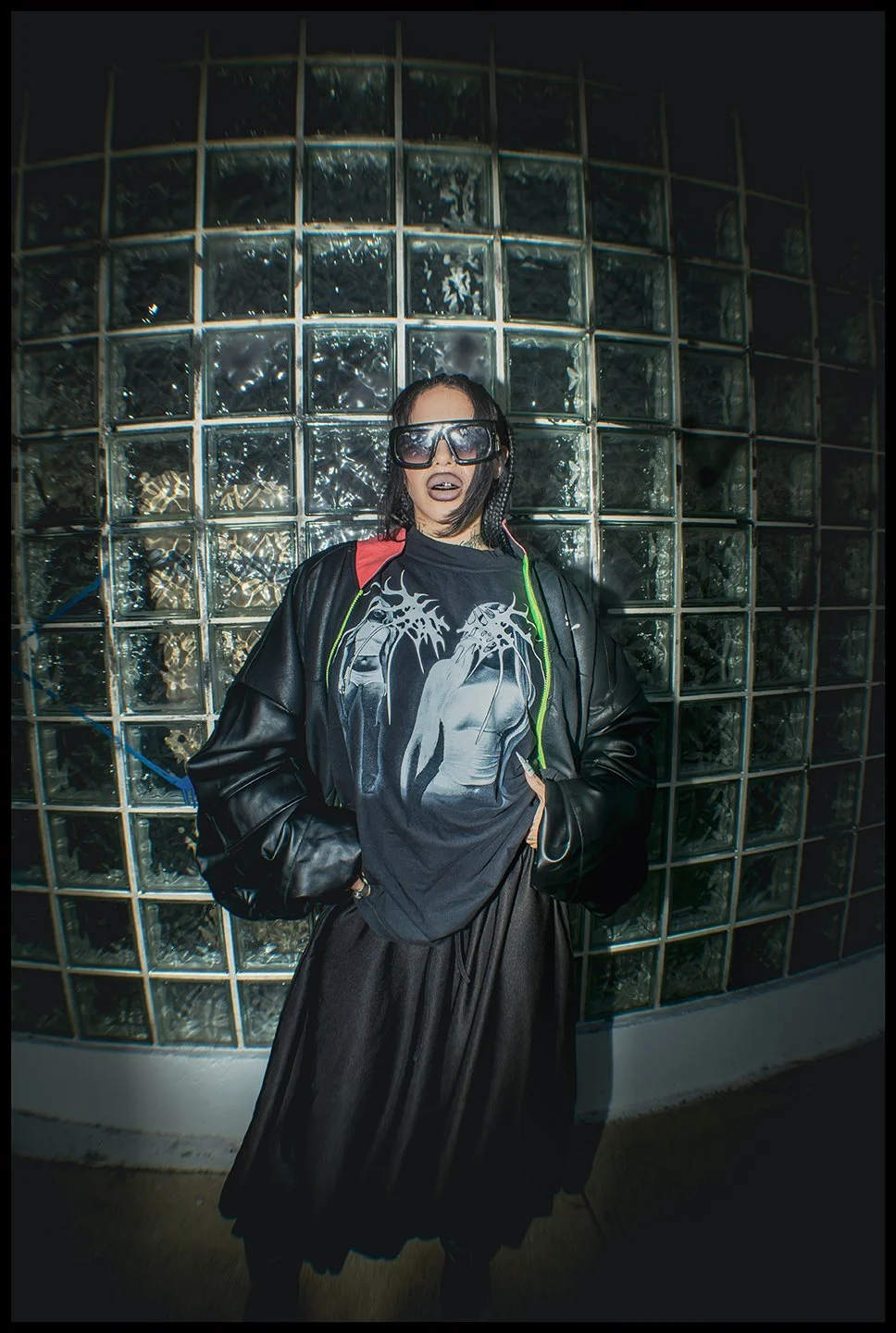 A woman standing in front of a glass block wall, wearing large sunglasses, a black graphic t-shirt, a black leather jacket with green lining, and a black skirt.