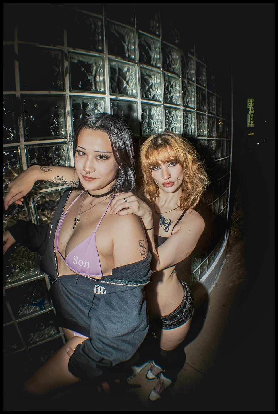 Two young women posing in front of a glass block wall at night, wearing revealing outfits with tattoos visible on their arms and shoulders.