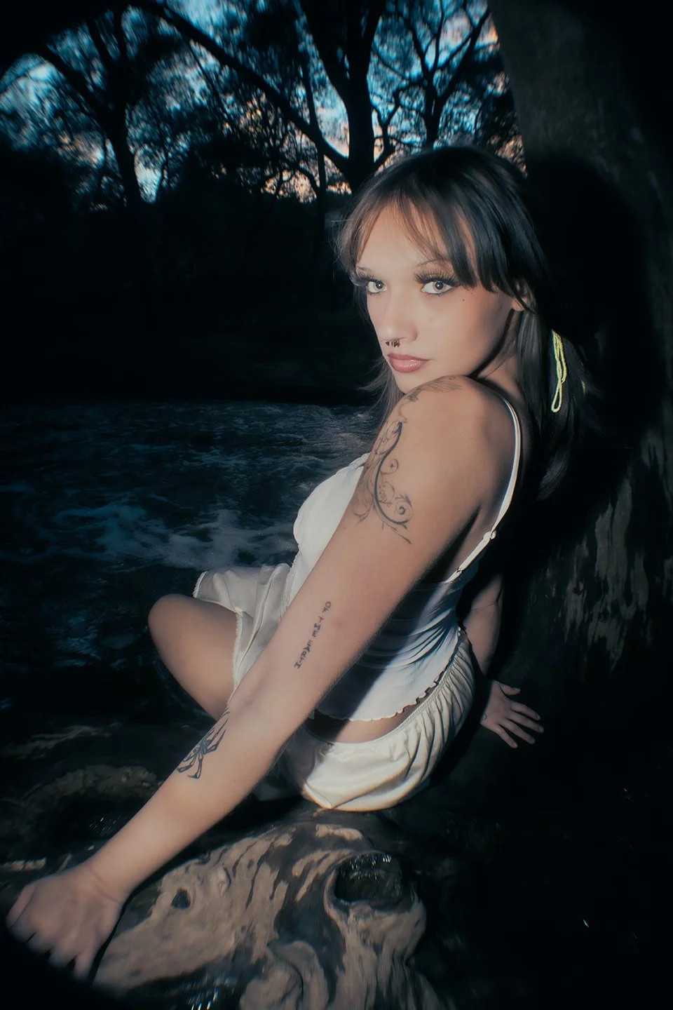 Young woman with tattoos and piercing, sitting in a river at dusk with trees in the background.