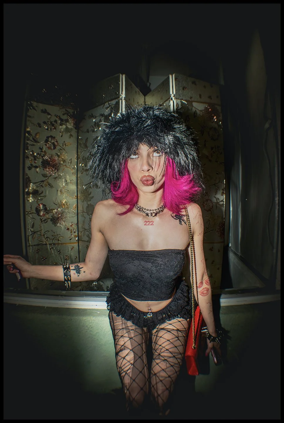 Person with pink hair wearing a black feathered hat, black strapless top, fishnet stockings, and jewelry, standing in front of decorative floral wallpaper.