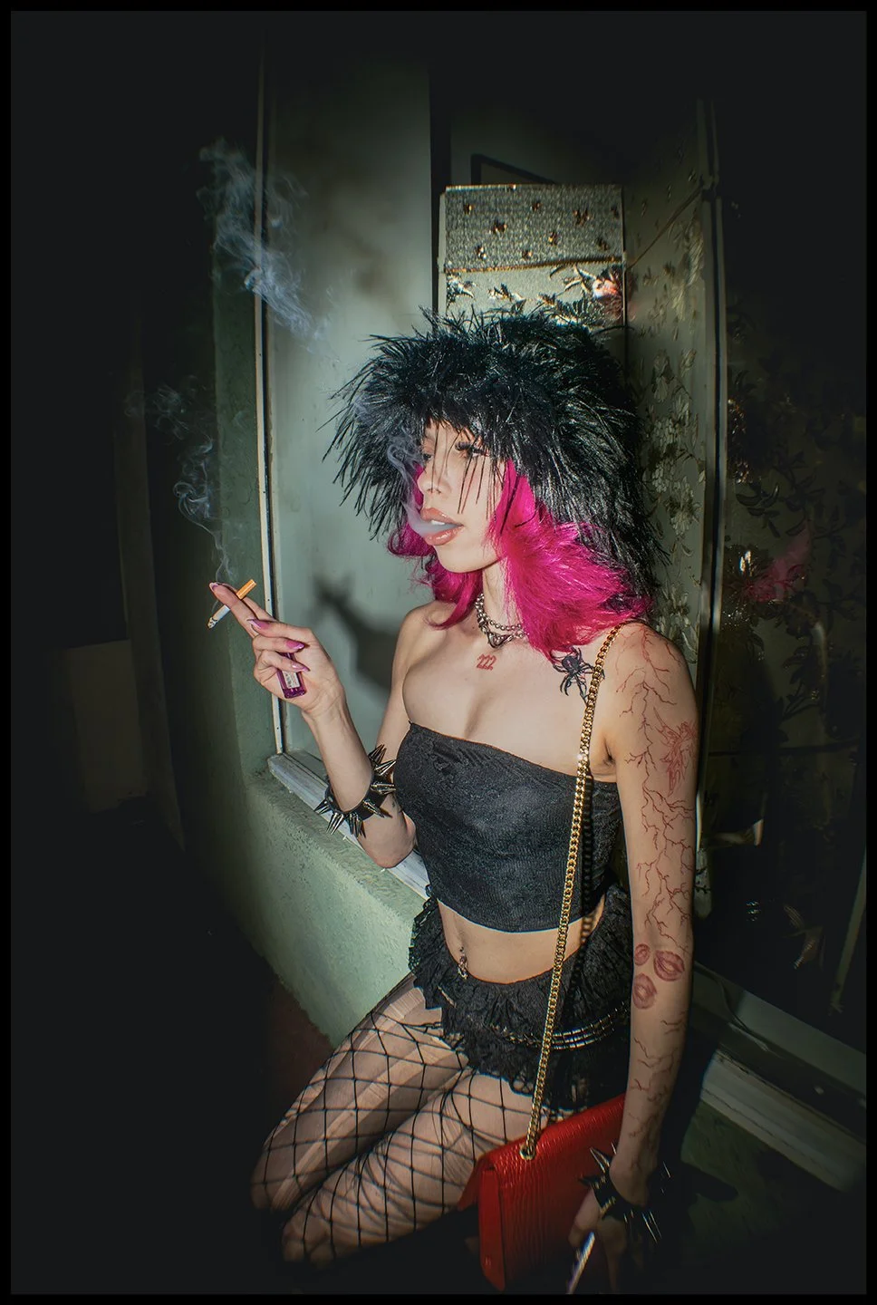 A woman with pink hair, black feathered hat, and tattoos, wearing a black strapless crop top and fishnet stockings, holding a cigarette and a red purse, in an indoor setting.