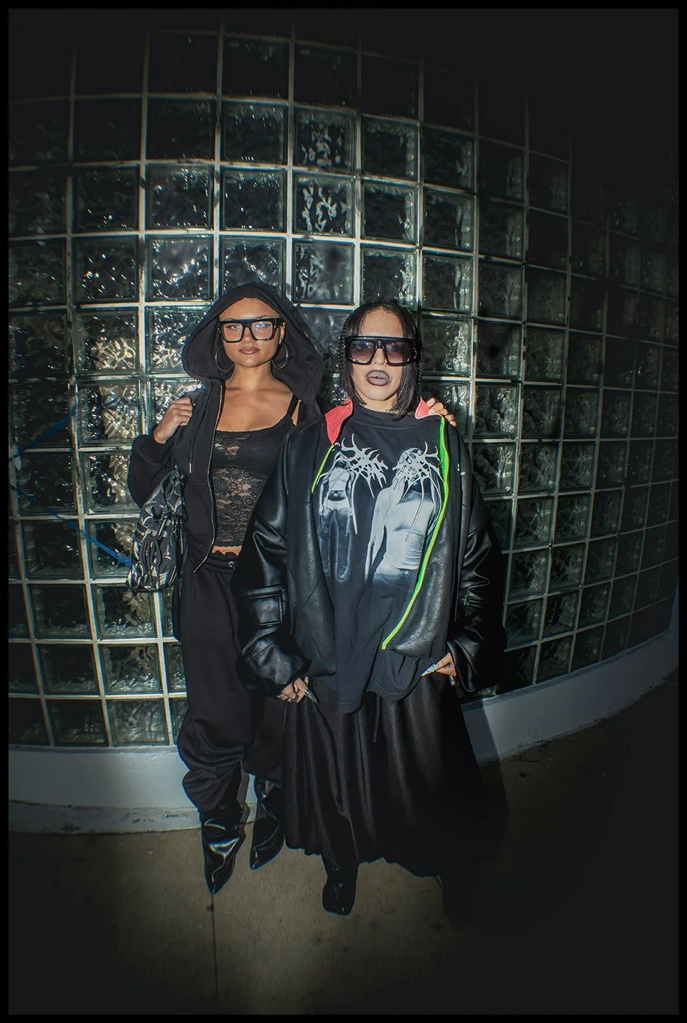 Two women standing in front of a glass block wall, both wearing black outfits and large glasses, with one woman wearing a graphic t-shirt and the other a hoodie.