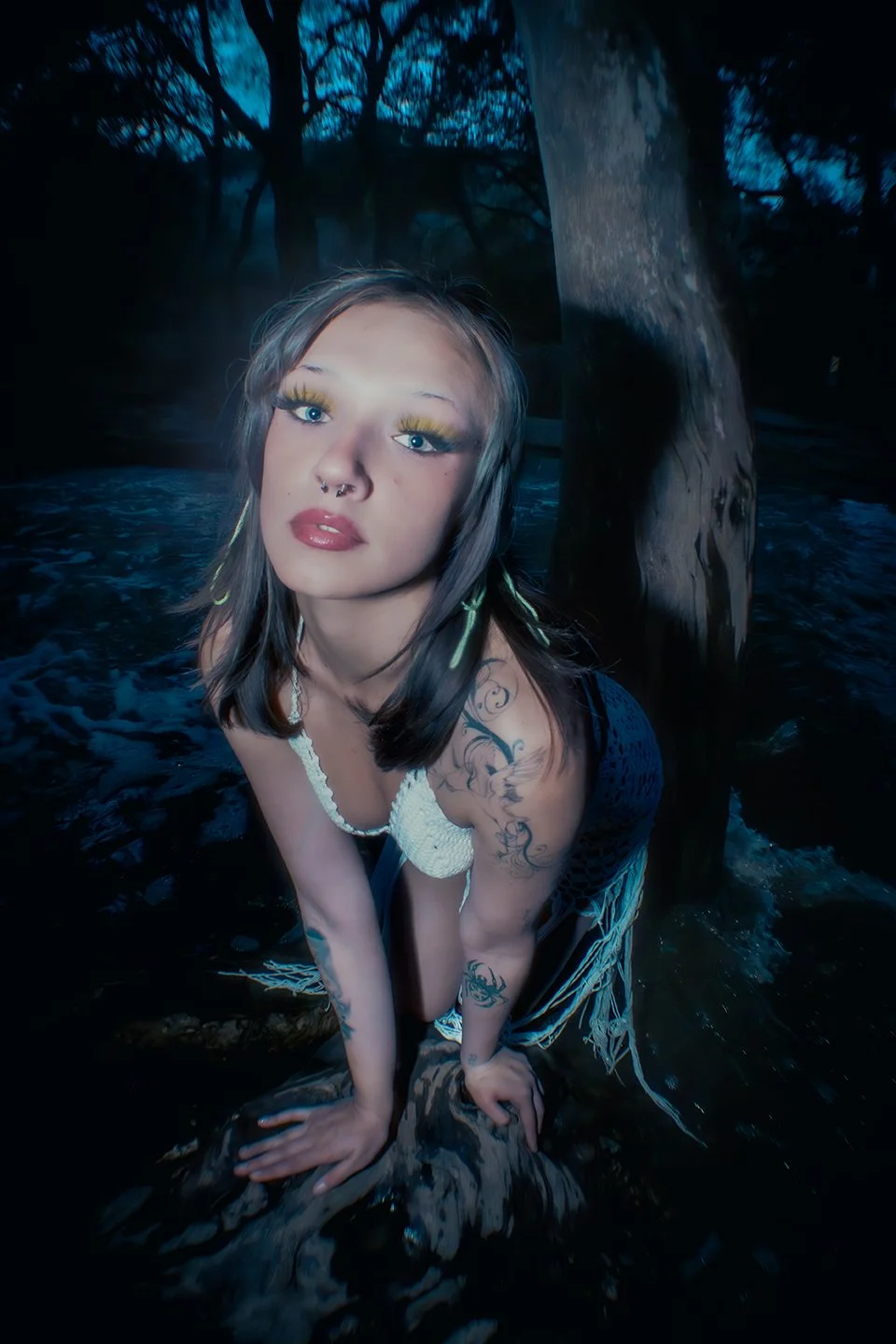 A young woman with tattoos and bright makeup crouches on a tree root in a dark outdoor setting with trees and water in the background.