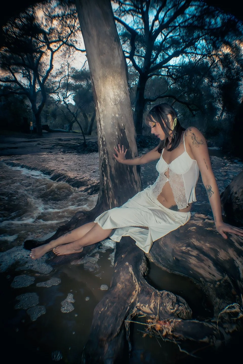 A woman in a white dress sitting on a large tree root in a stream, touching a tree trunk surrounded by trees with blue sky visible through the branches.