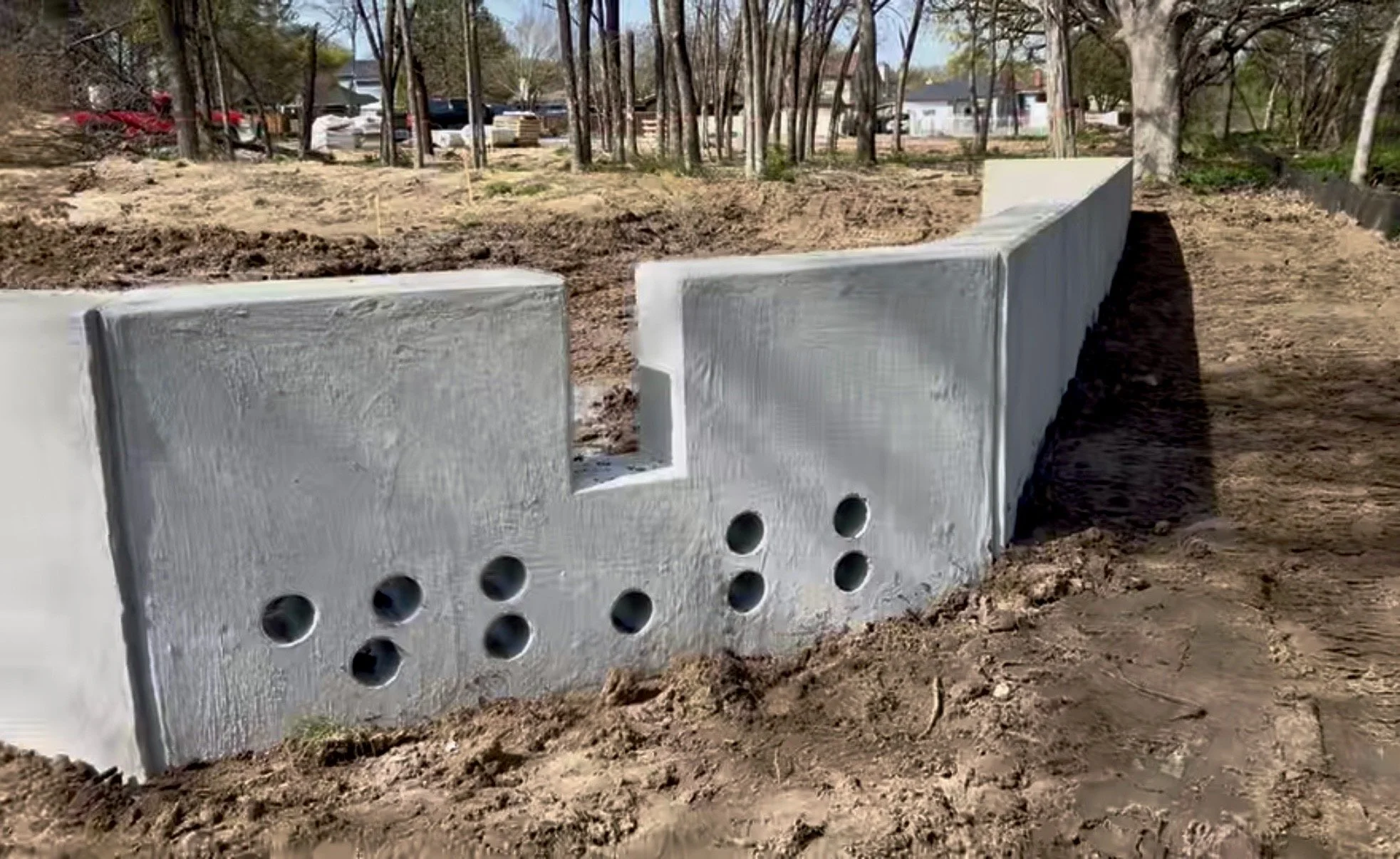 A concrete wall under construction with black drainage pipe visible beneath it, in a residential neighborhood with large houses and manicured lawns.