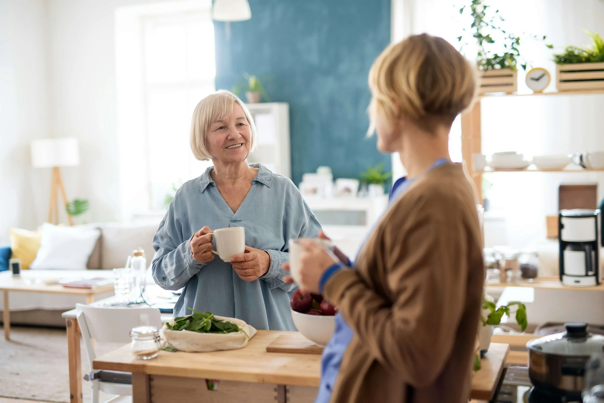 A mother and daughter stand in a kitchen, looking at each other while holding coffee mugs