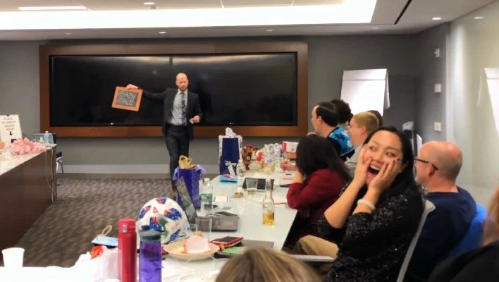 A man in a suit standing in front of a large black screen, giving a presentation to a group of people sitting at a long table in a conference room. The group is engaged, with one woman smiling and covering her mouth.