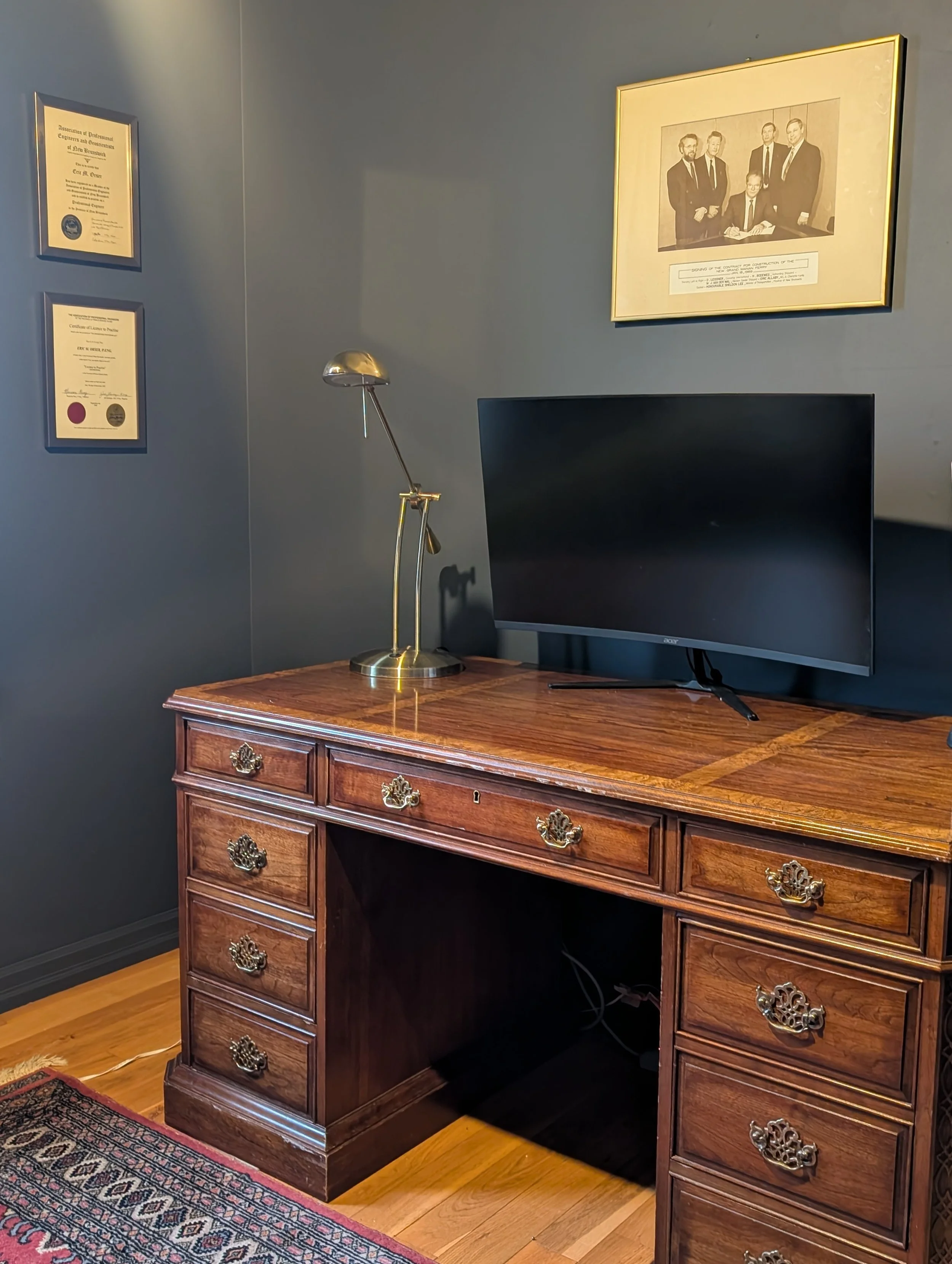 A wooden desk with brass ornate handles, a black computer monitor, a gold desk lamp, and framed certificates and a black-and-white photograph on a blue wall.