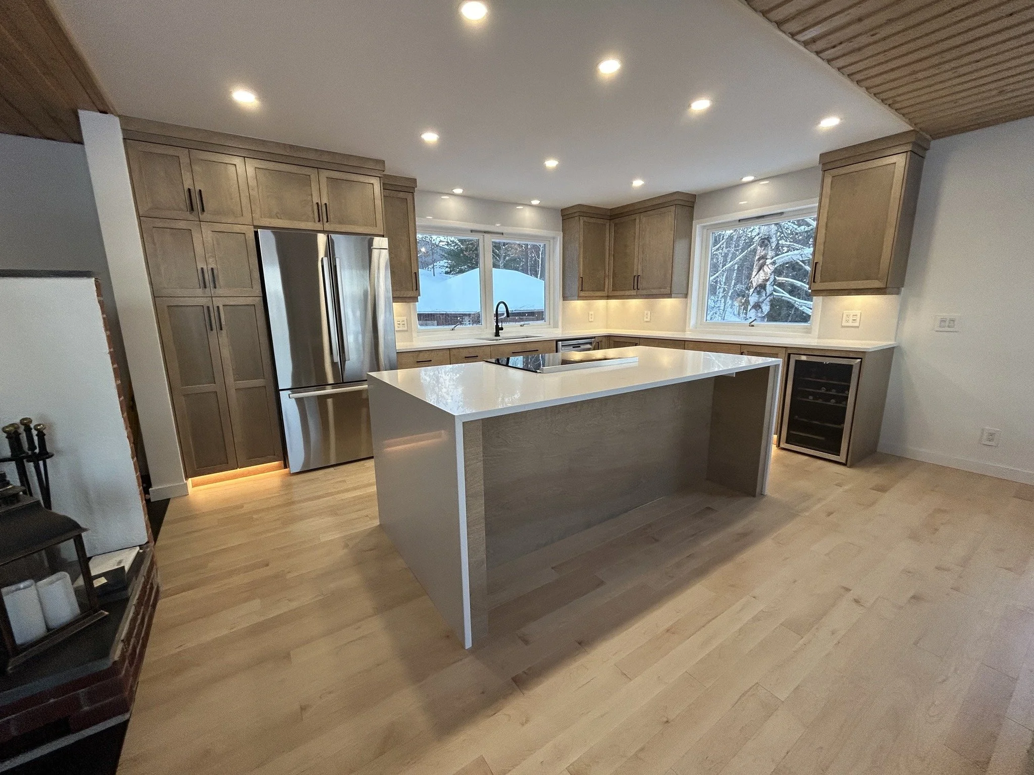Modern kitchen with wooden cabinets, stainless steel refrigerator, white island, and large windows showing snow-covered trees outside.