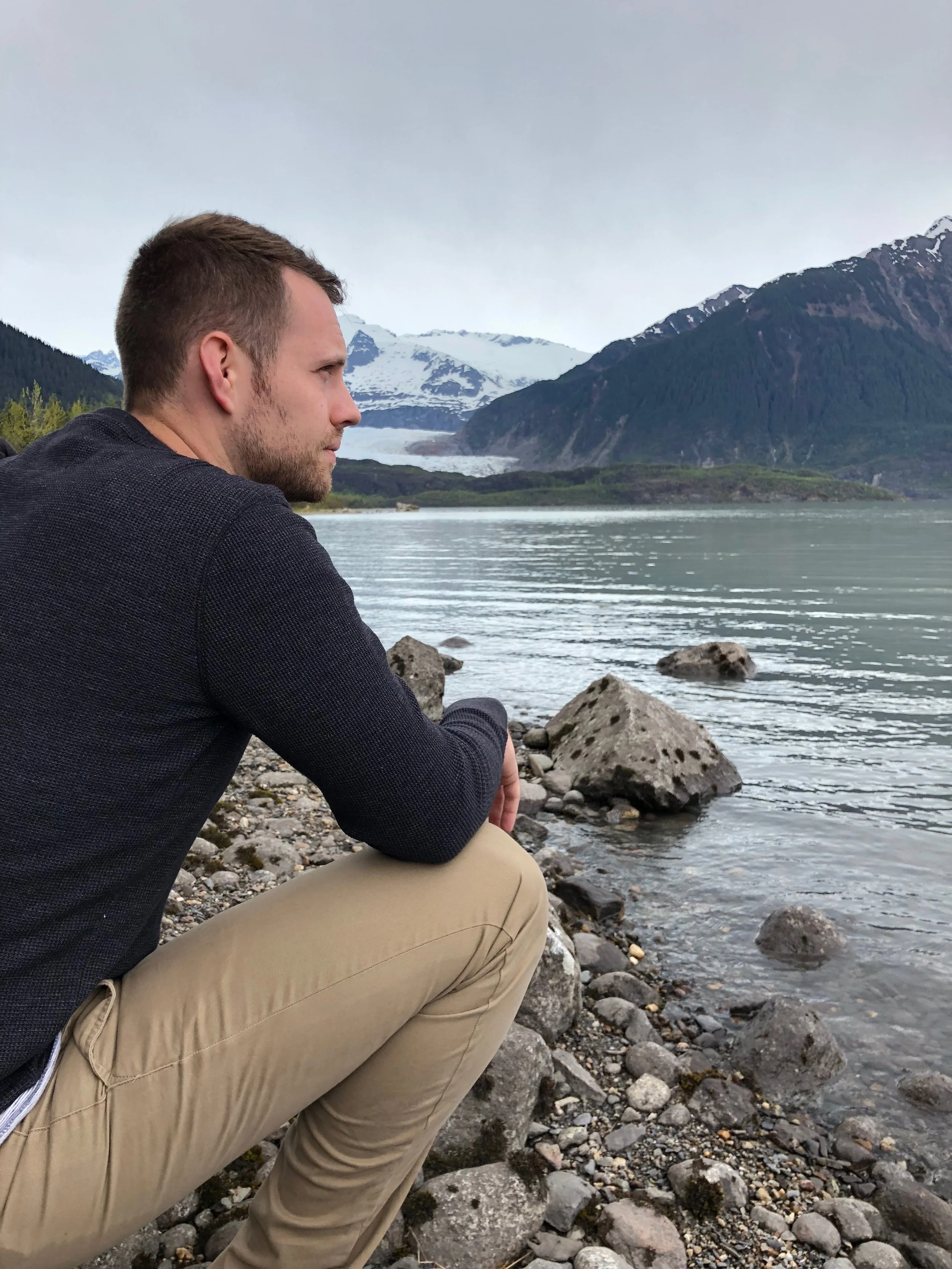 A man, Tyler Braun, crouching on a rocky shoreline at the Mendenhall Glacier in Alaska, looking at a lake with mountains and snow-capped peaks in the background.