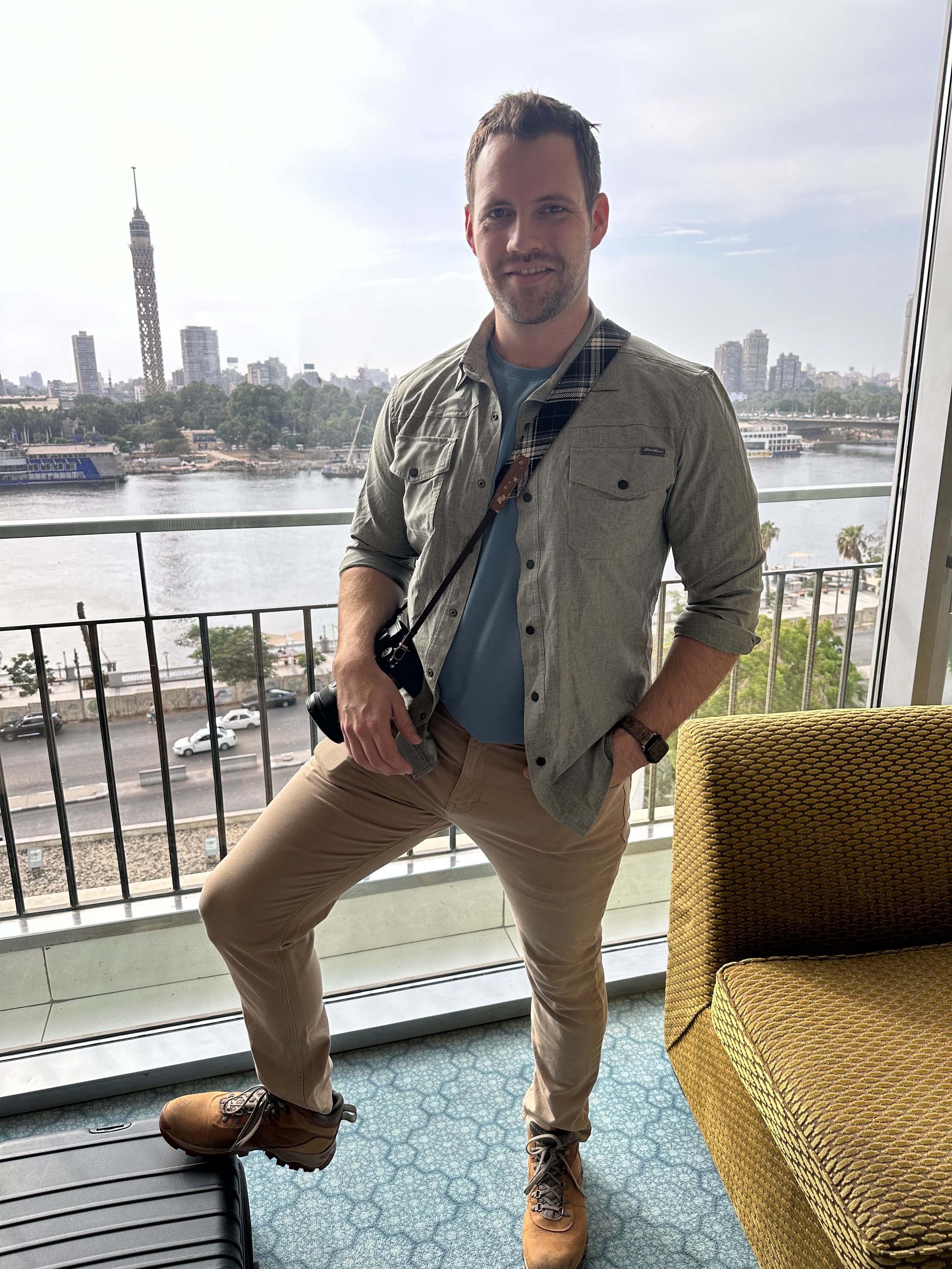 A man, Tyler Braun, standing on a balcony overlooking the Cairo skyline and Nile River, wearing a casual outfit with a camera hanging from his neck.