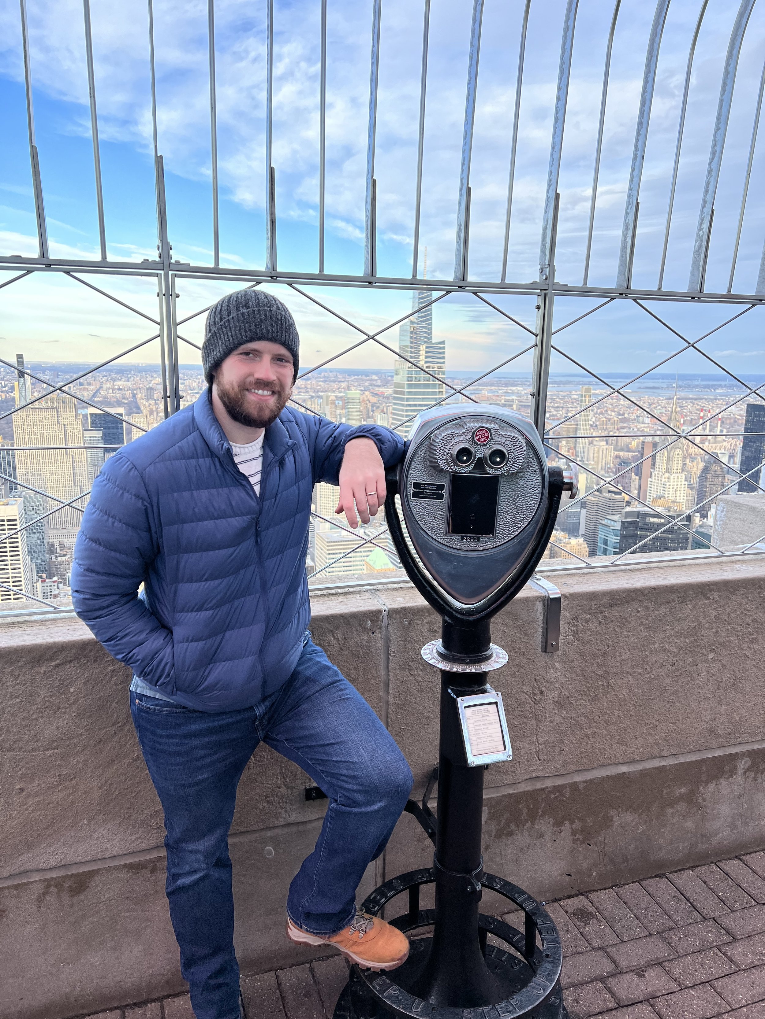 Tyler Braun at the top of the Empire State Building with a beard wearing a black beanie, a blue puffer jacket, and jeans, standing on a rooftop with a cityscape of New York City view behind him, next to a coin-operated binocular viewer.