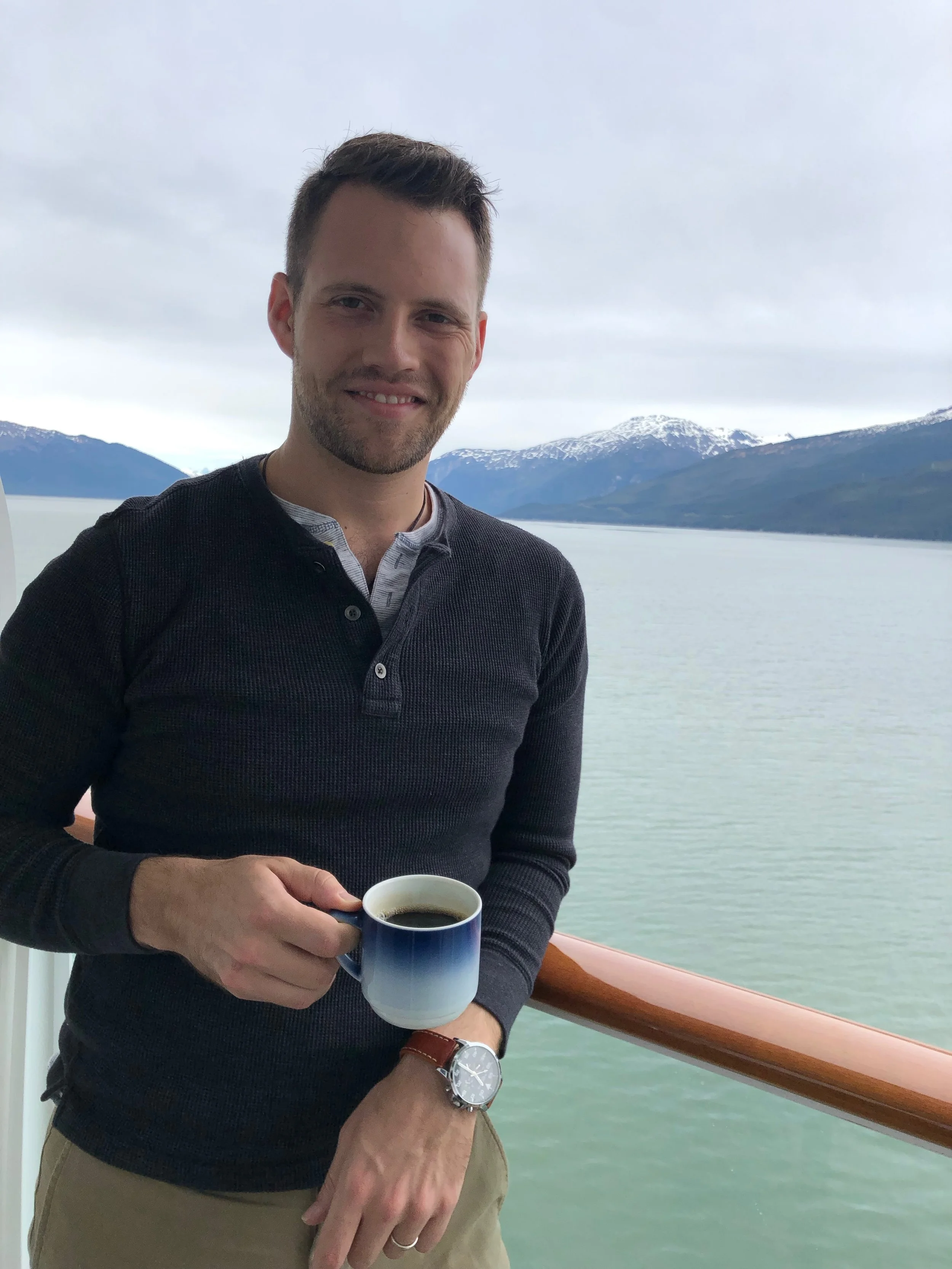 A man, Tyler Braun, holding a cup of coffee on a ship in Alaska, with a body of water and snow-capped mountains in the background.