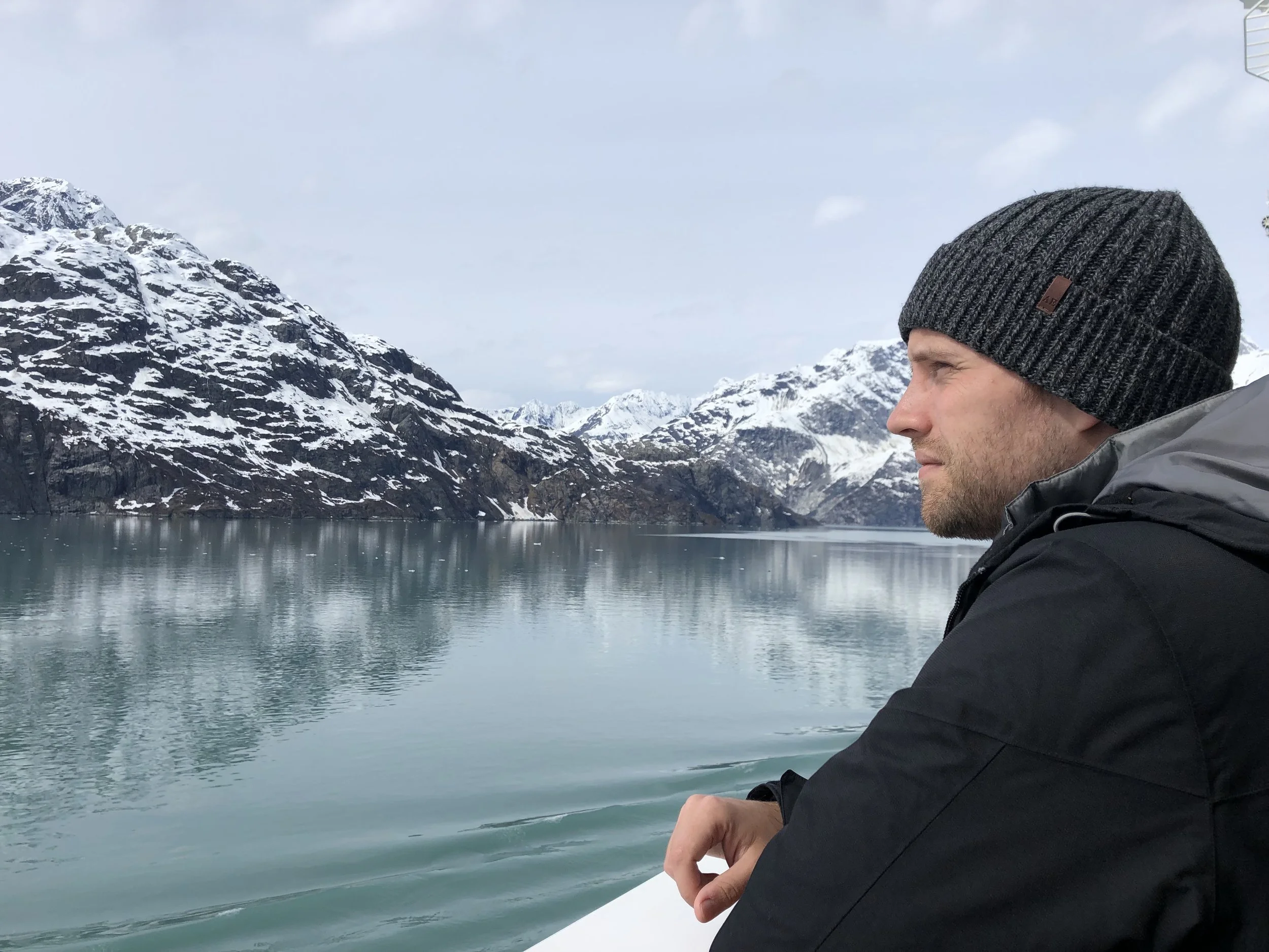 A man, Tyler Braun, wearing a black beanie and jacket looking at a snowy mountain landscape across a calm body of water in Glacier Bay in Alaska.
