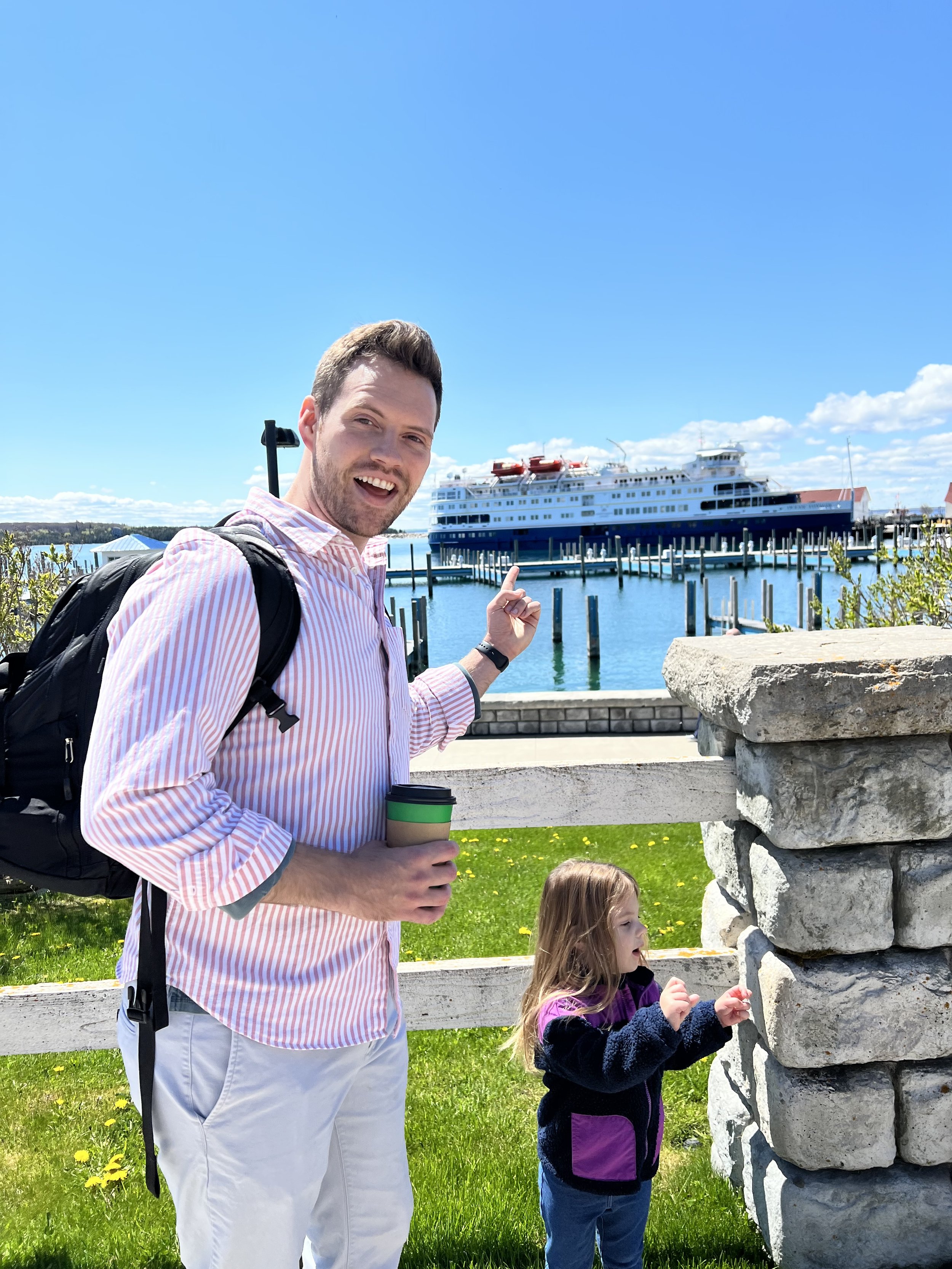 A man, Tyler Braun, with a backpack holding a coffee cup and smiling, pointing at a boat docked at a marina, with a young girl standing nearby looking at a stone wall, on a sunny day with blue sky and scattered clouds in Mackinac Island Michigan.