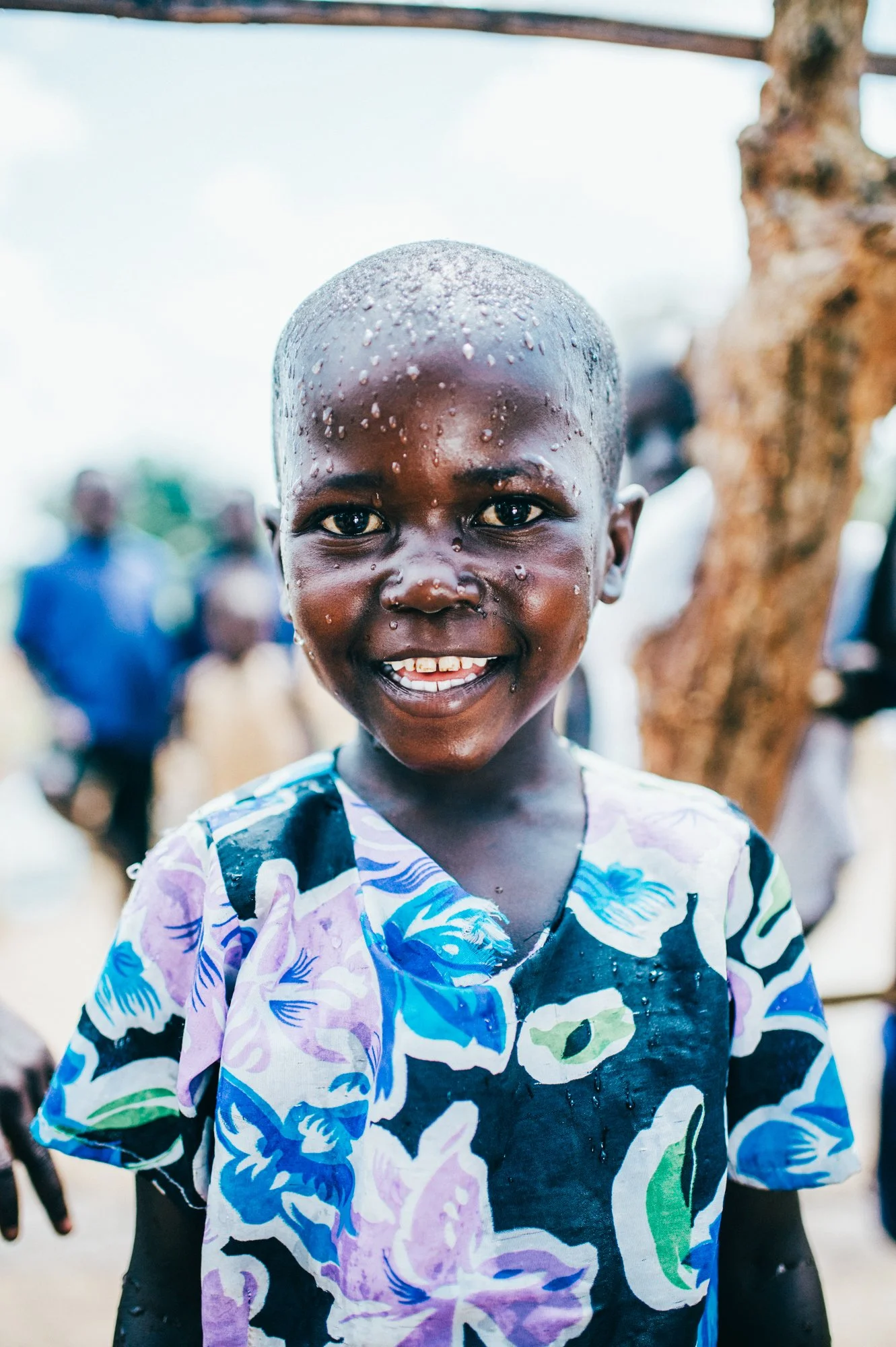 Thirst Project - A young boy with short hair and a big smile, wet from water, wearing a floral shirt, standing outdoors with a tree and blurry people in the background.