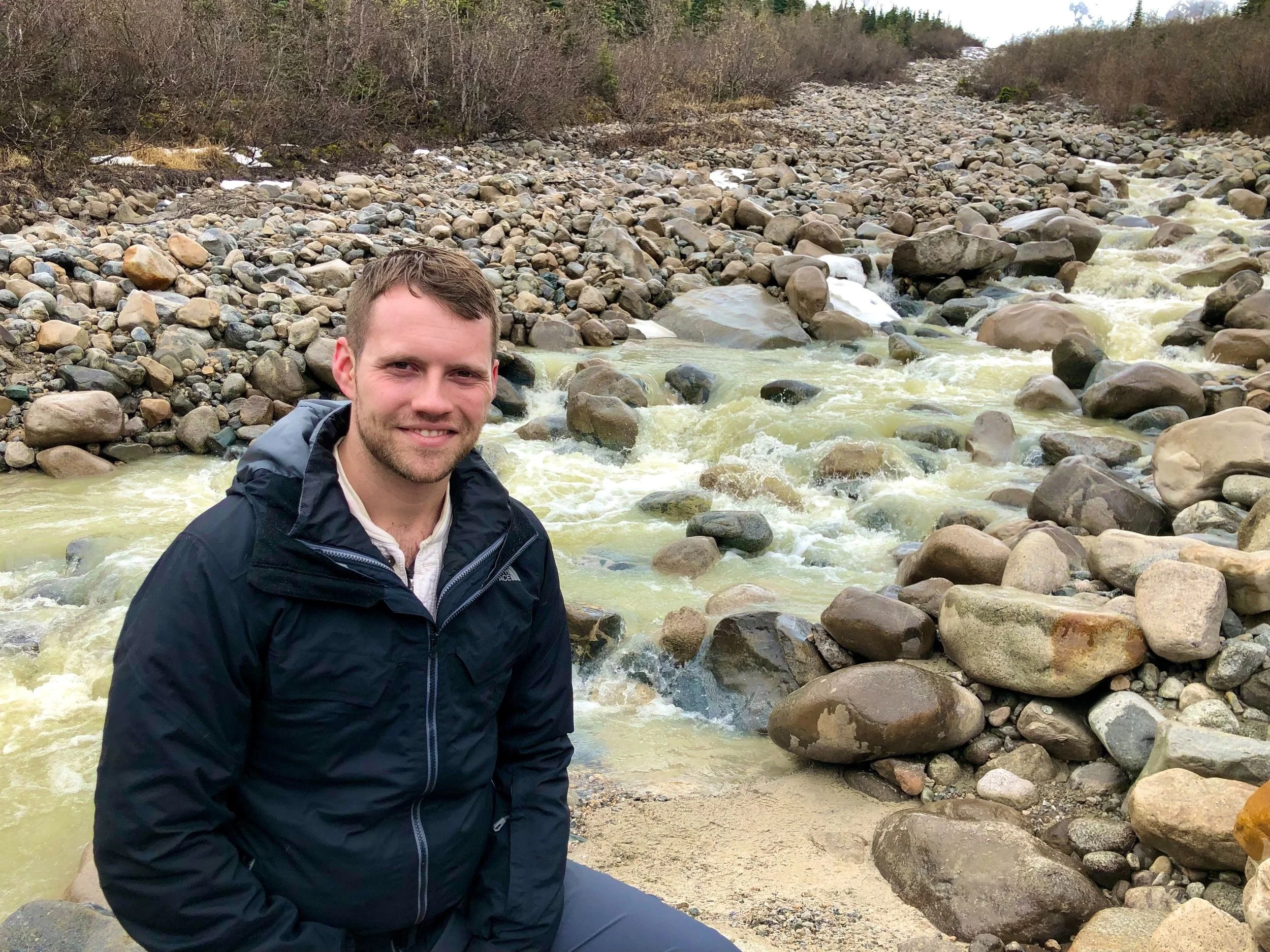 A man, Tyler Braun, in Alaska in a dark jacket sits on the rocks beside a flowing mountain stream with a wooded hillside in the background.