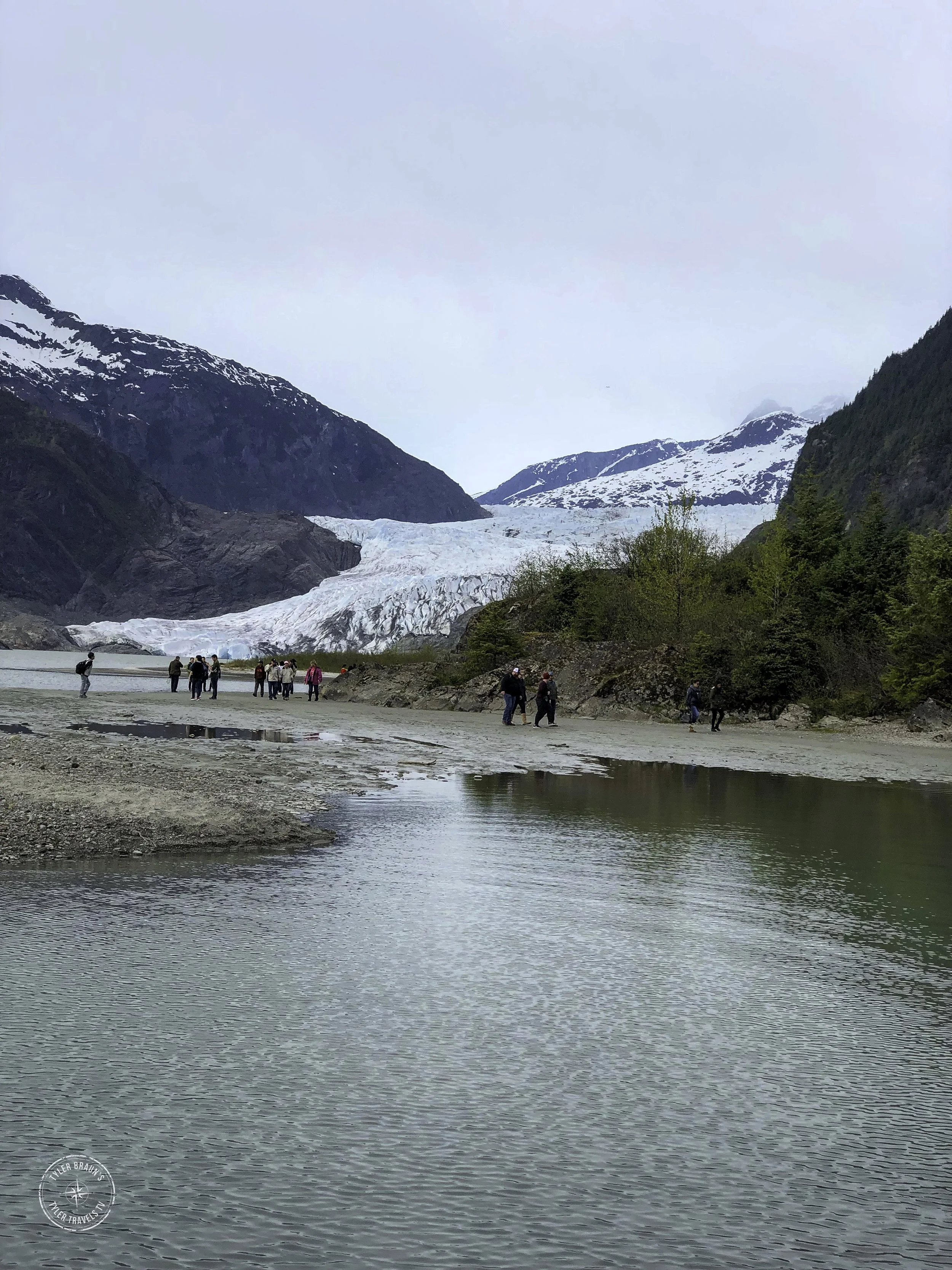A group of people walks along the shore of the Mendenhall Glacier in a mountainous area, with snow on the peaks and green trees on the slopes.