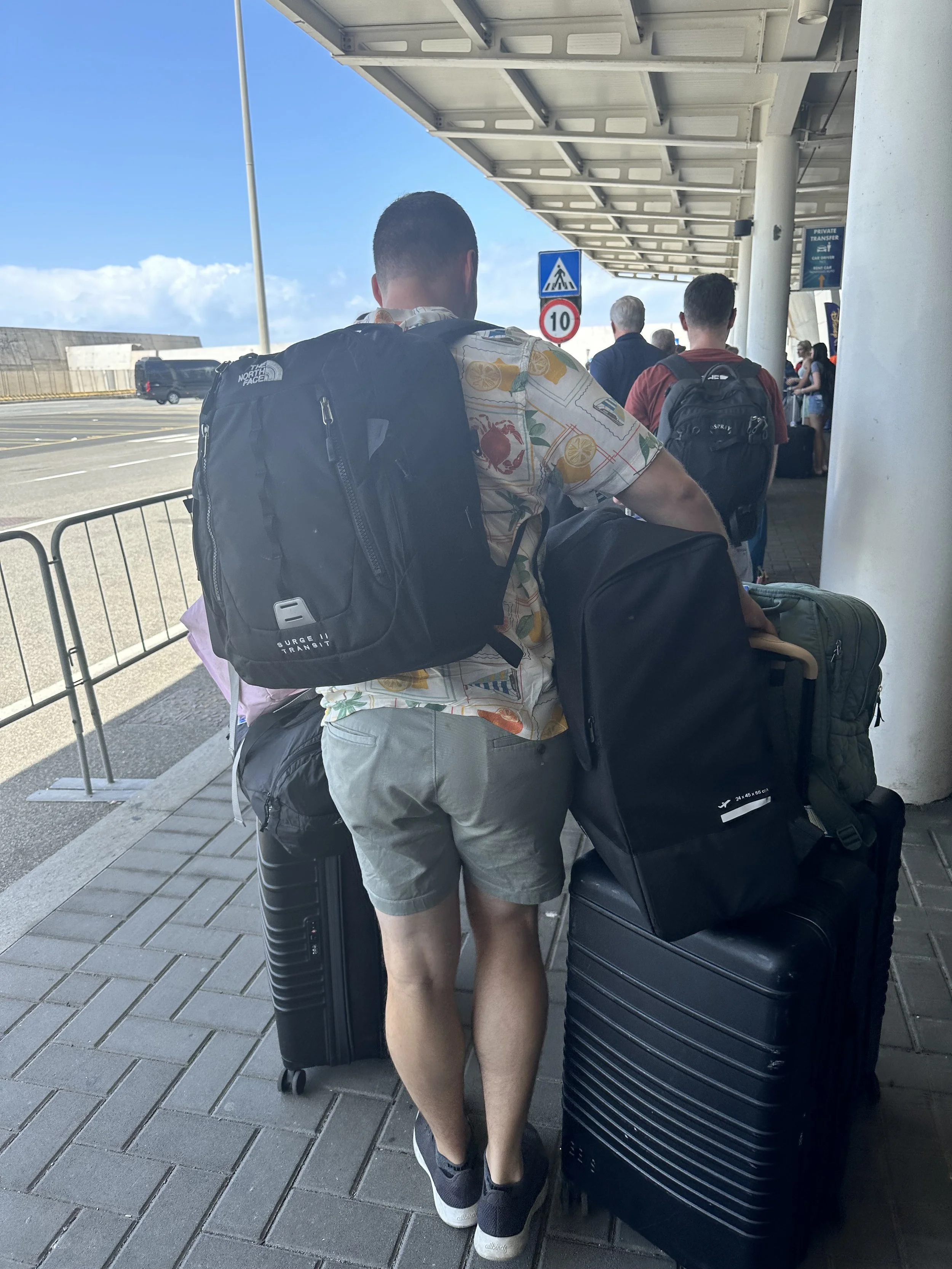 People standing in line at an airport terminal, with luggage and backpacks, under a covered walkway.