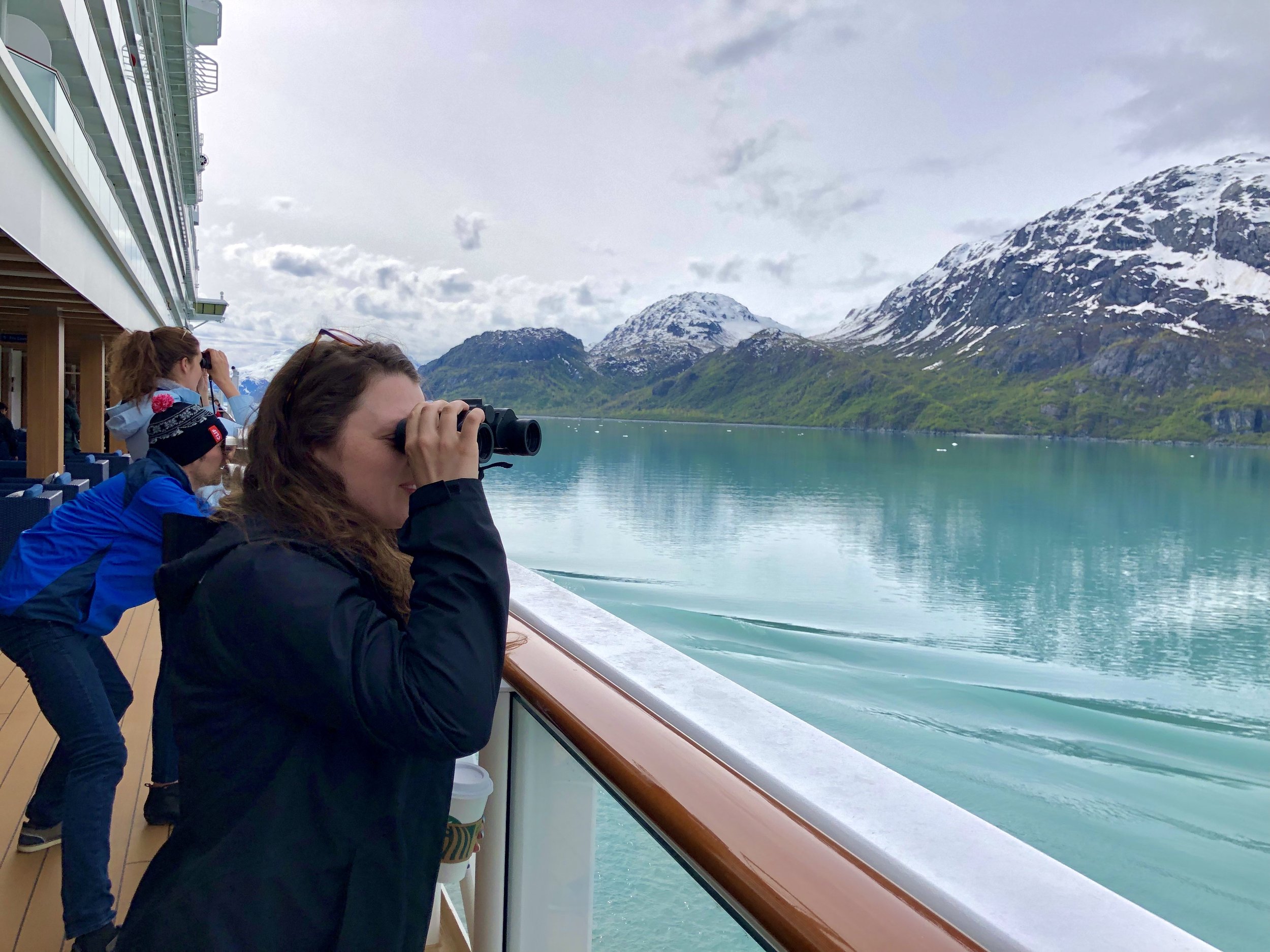 Multiple people on a cruise ship deck looking out and taking photos of icy mountains and calm turquoise water in Alaska.