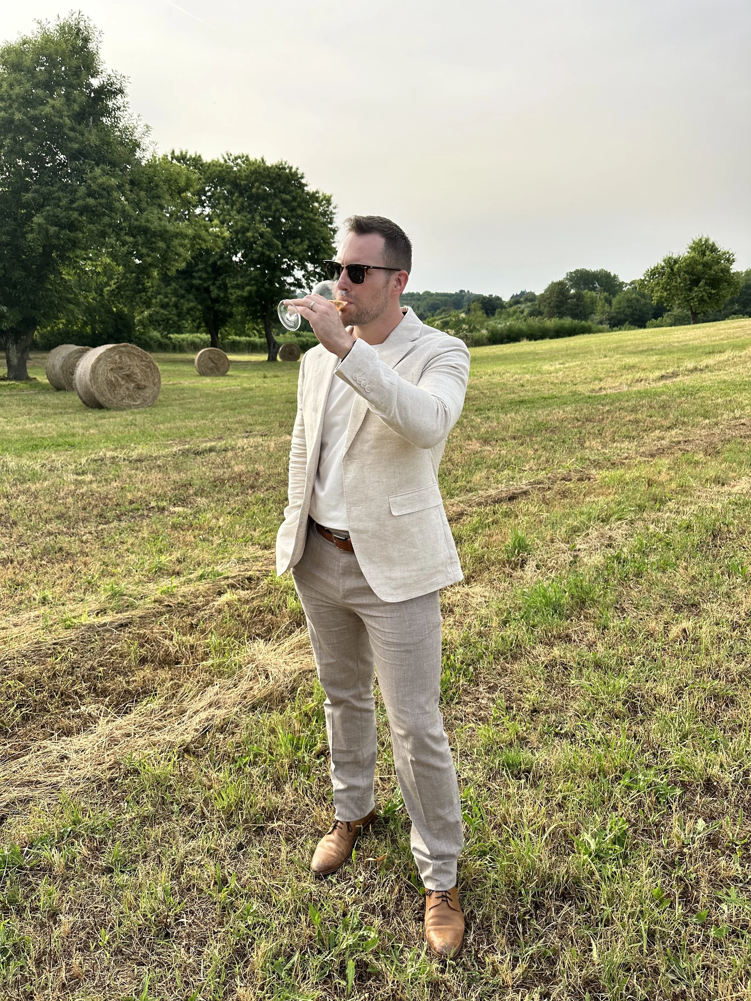 A man, Tyler Braun, wearing a light-colored suit and sunglasses is standing in a grassy field, drinking from a glass of wine, with hay bales and trees in the background in Italy.