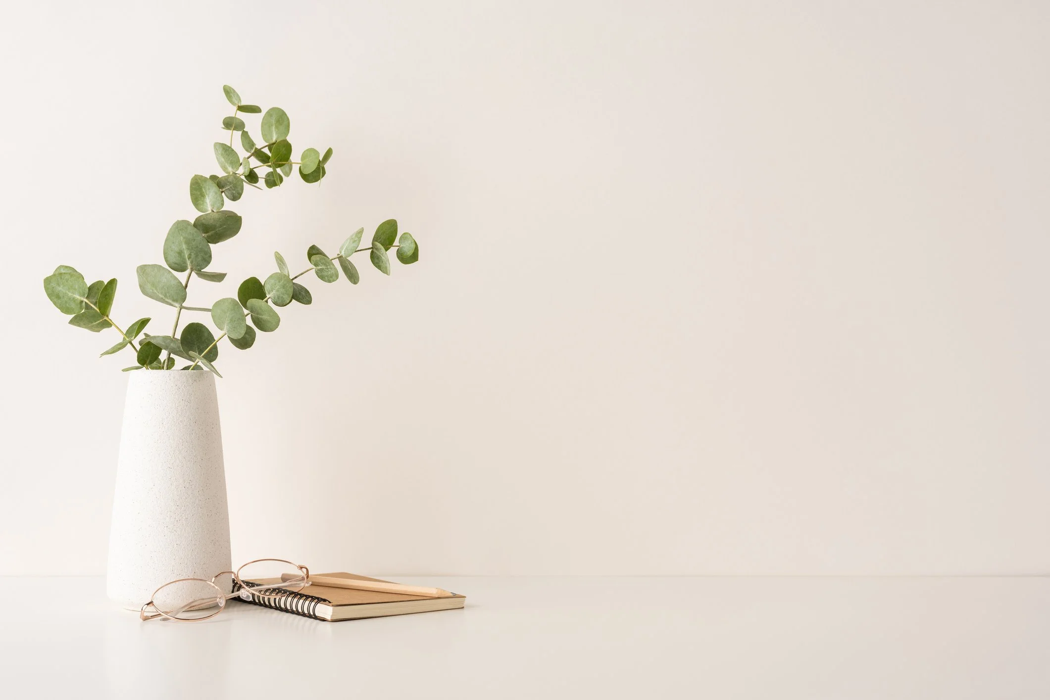 A white vase with green eucalyptus branches on a minimalist desk, accompanied by a notebook, glasses, and a pen.