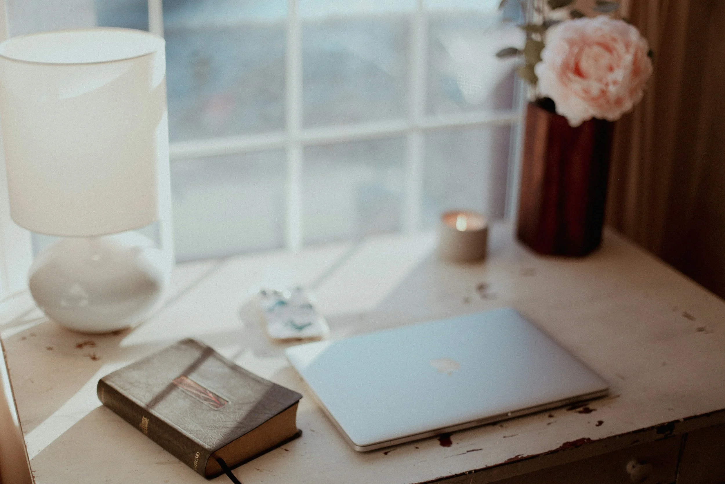 sunlit window with an apple macbook closed on top of a light wooden desk with a brown book on top.