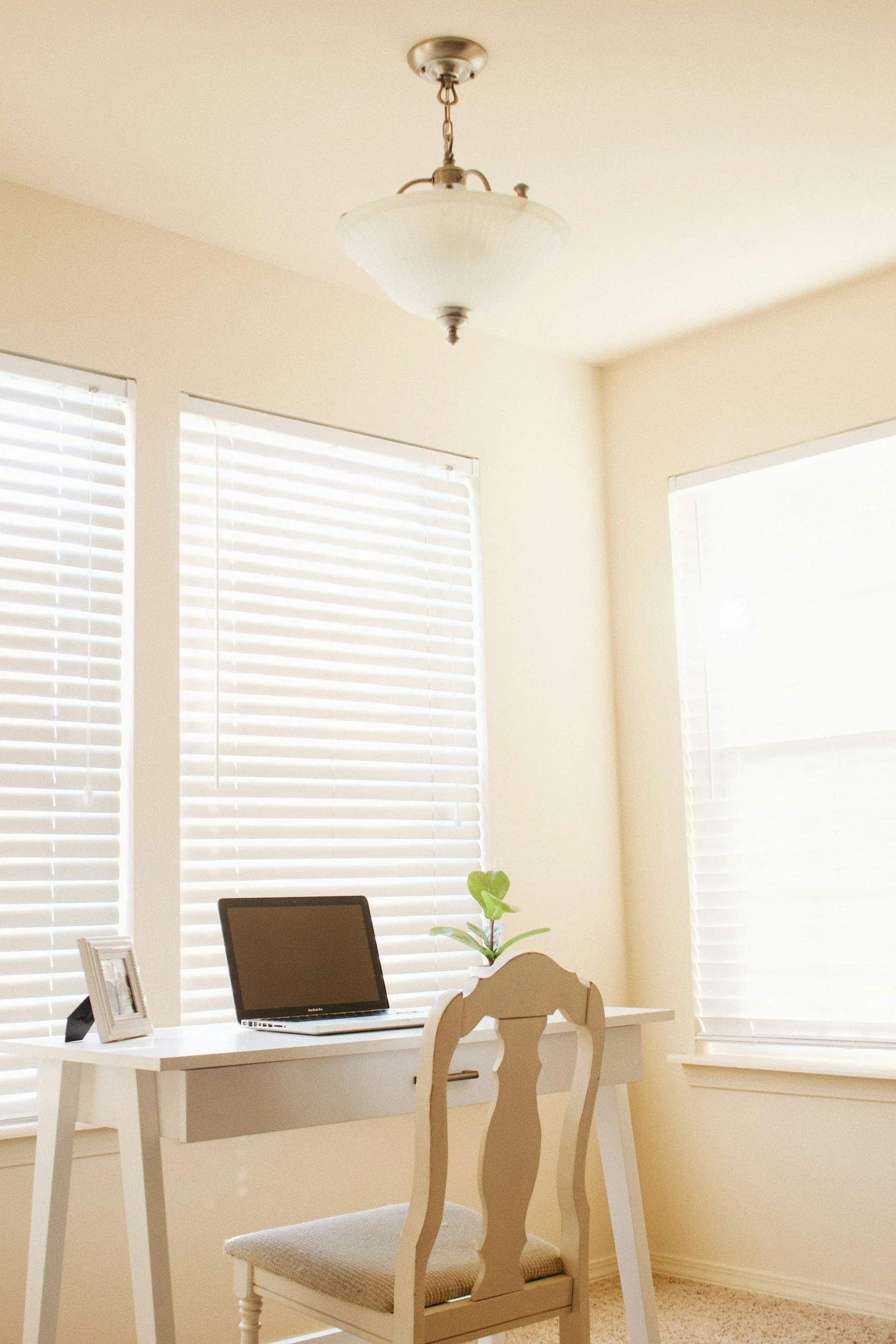 Home office with white desk, laptop, framed photo, and small plant near large windows with white blinds, beige walls, and ceiling light fixture.