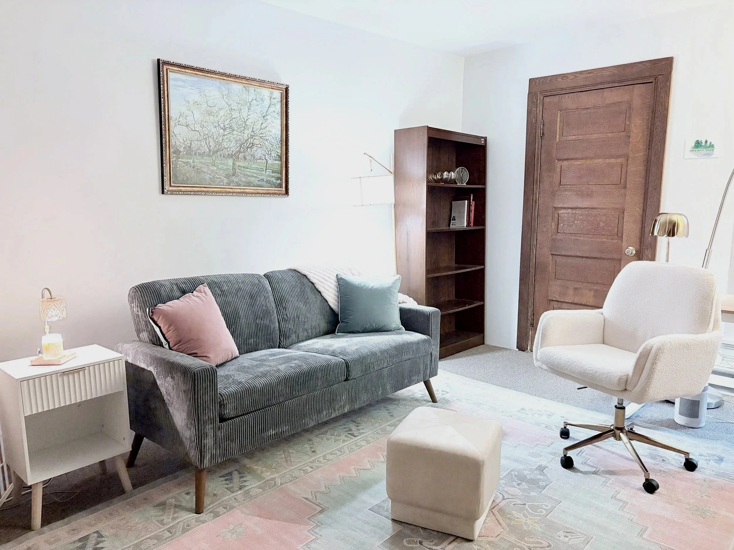 A cozy living room with a gray-striped sofa, pastel-colored pillows, a white side table with a lamp, a wooden bookshelf with decorative glasses and books, a large framed painting of apple trees on the wall, a wooden door, a beige office chair, a white ottoman, a floor lamp, and a soft pastel rug.