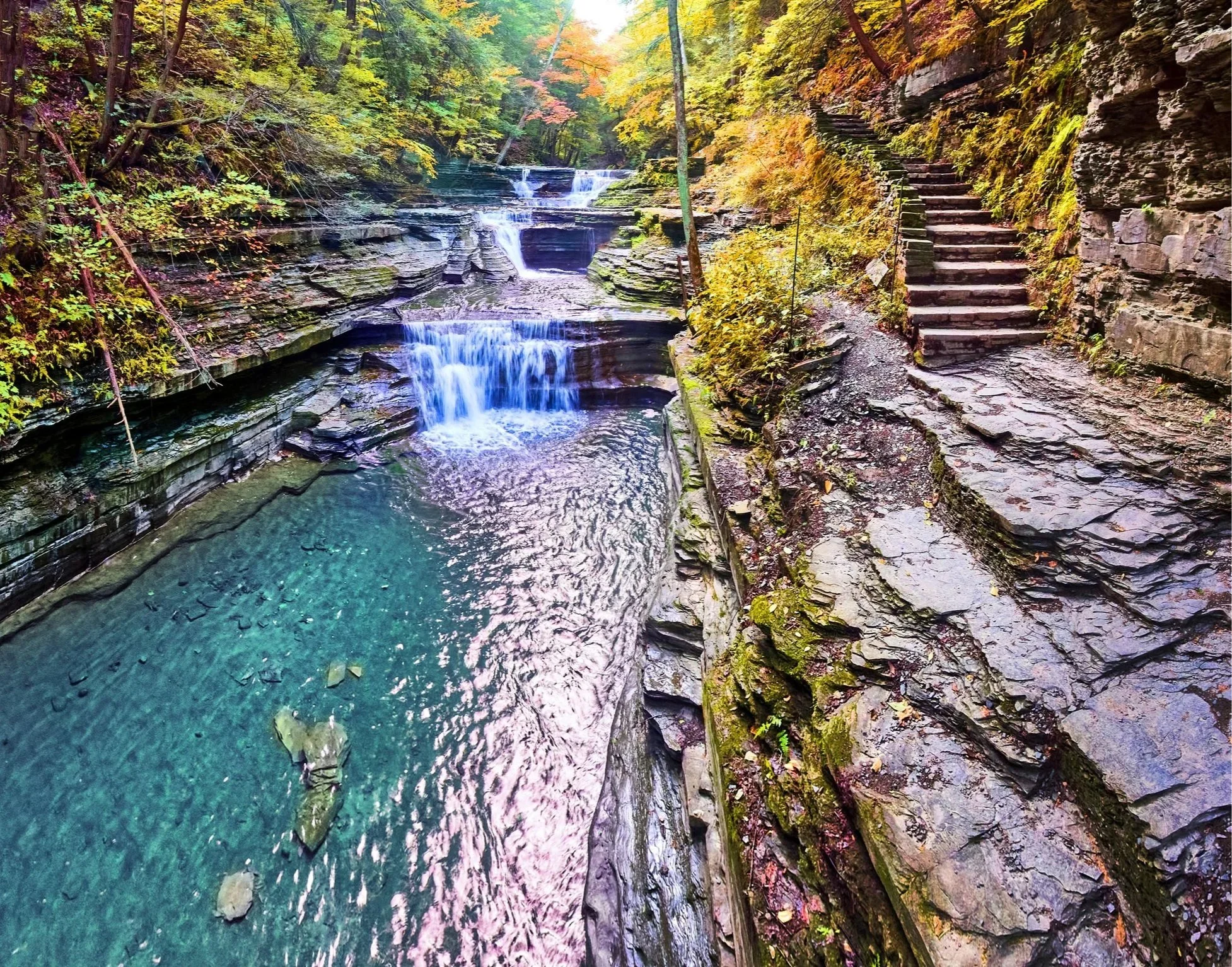 A scenic river with clear water flowing over rocks, surrounded by trees with autumn leaves, and a set of stone steps along a rocky cliff on the right.