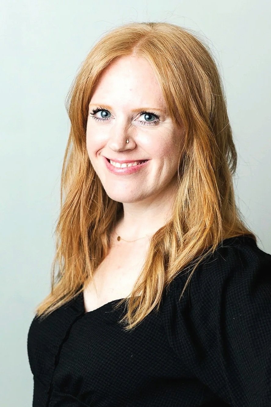 A woman with long red hair, blue eyes, and a nose piercing, smiling in front of a light-colored background.