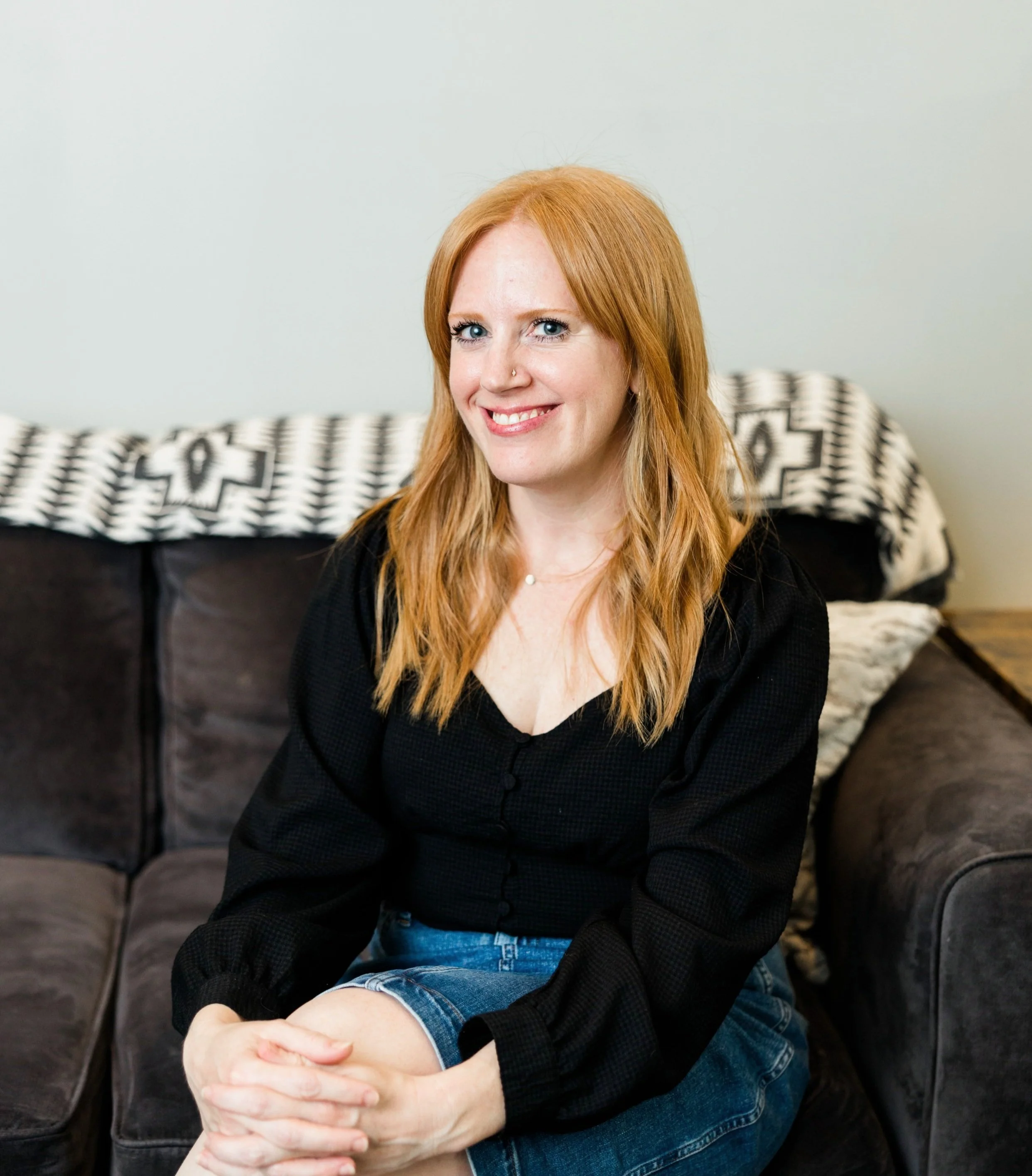 A woman with red hair, blue eyes, and a nose piercing smiling while sitting on a black leather couch with a patterned blanket in the background.