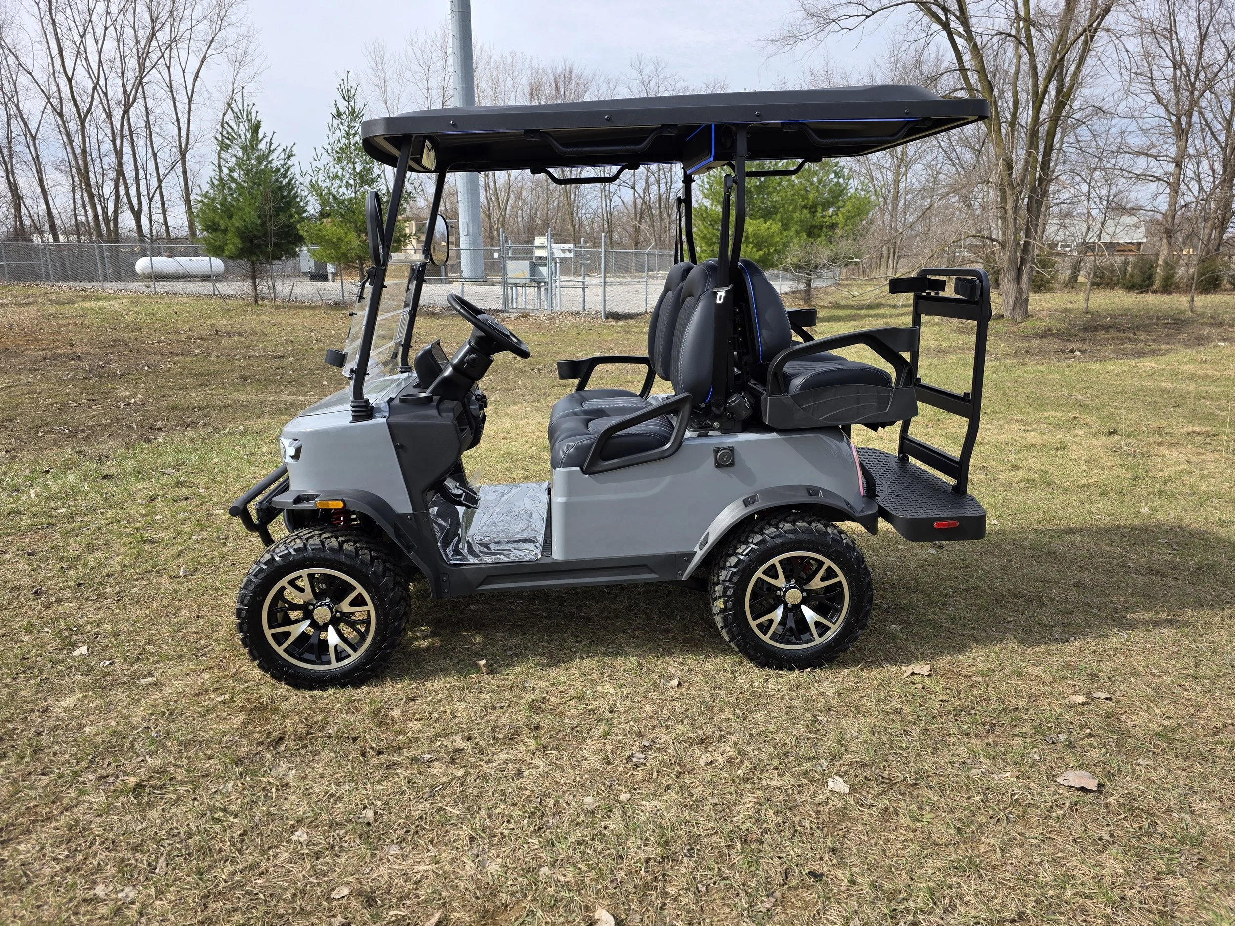 Gray golf cart with four black seats, black roof, and off-road tires, parked on a grassy area with trees and a chainlink fence in the background.